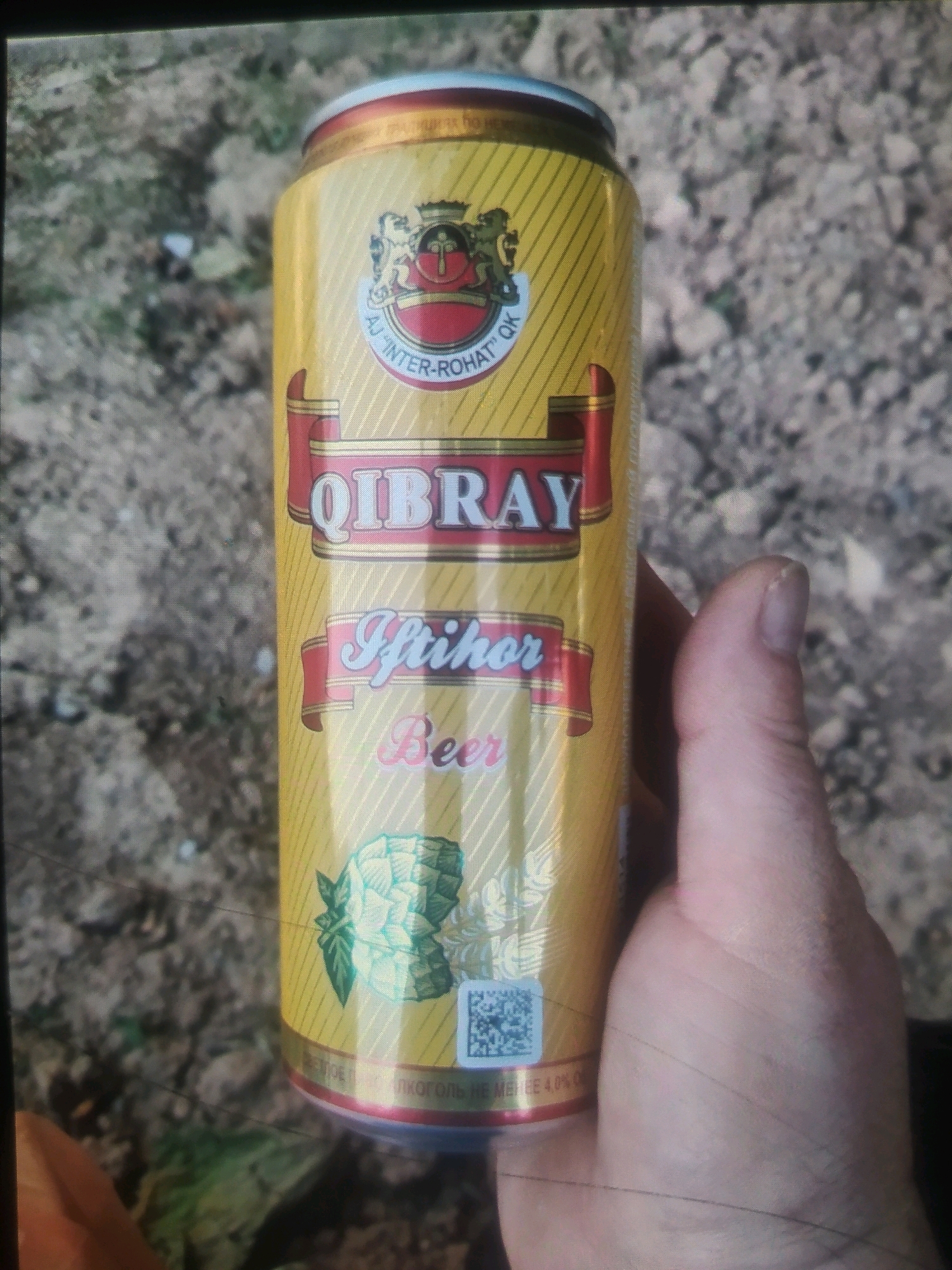 A close-up, eye-level shot captures a yellow beverage can with red and white labeling held in a person's right hand, positioned in the foreground. The can prominently displays the text "QIBRAY" in large, stylized white letters with a red outline. Below this, "Iftikhor" is written in a cursive script, followed by "Beer" in a similar font. A depiction of a hop cone with leaves is visible on the lower portion of the can, alongside a QR code. The background consists of dry, mottled ground, suggestive of an outdoor environment. The lighting appears to be natural, casting a slight shadow on the can. The details of the hand suggest it belongs to an adult. The context implies the can is a product of Birlik, Uzbekistan, as indicated by the presence of text in Cyrillic script on the lower rim of the can, likely related to its origin or composition.