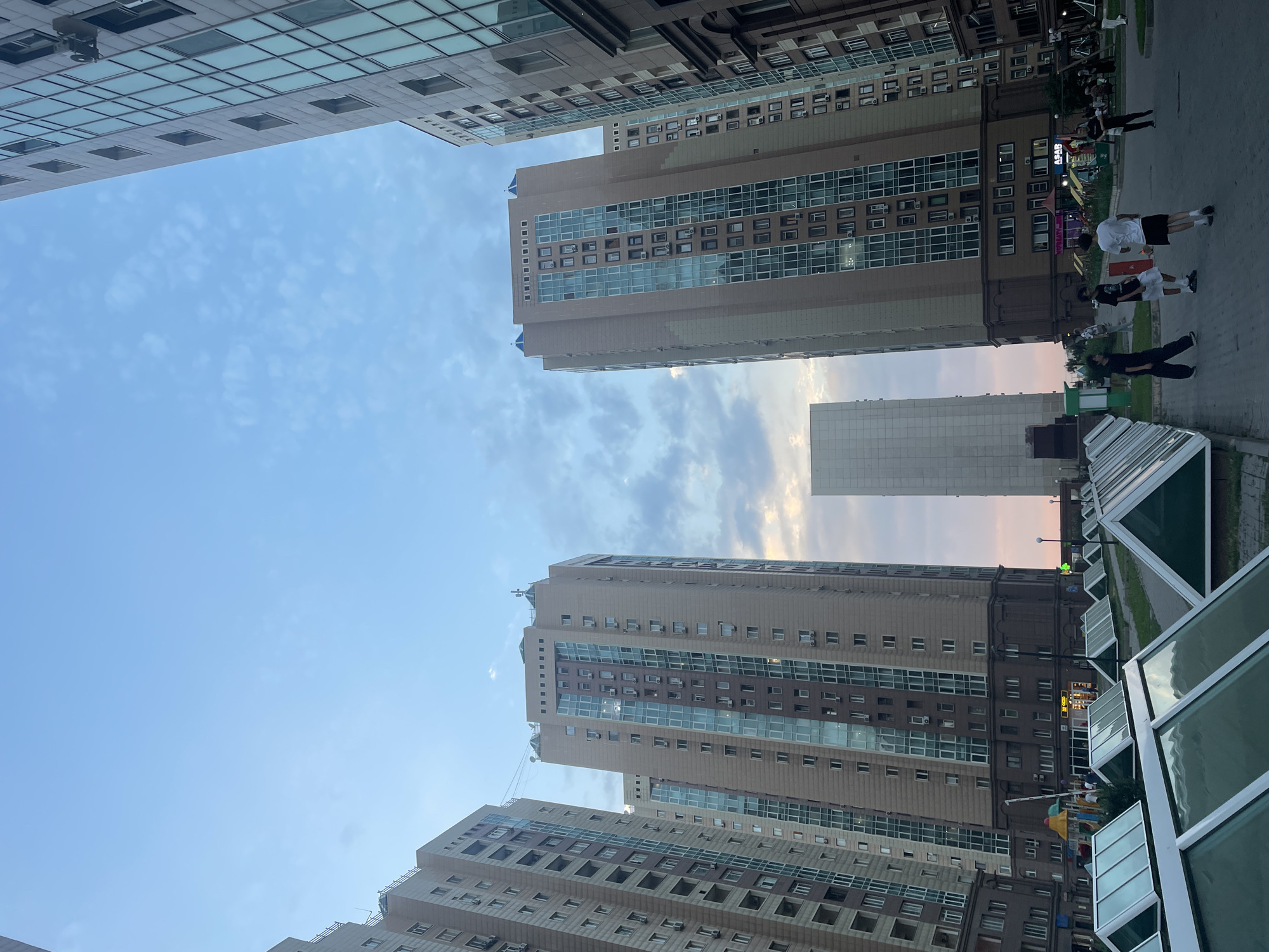 The image captures a view of several modern, tall residential buildings in the city of Almaty, Kazakhstan. The buildings are characterized by their contemporary architecture, featuring large glass windows and varying facade materials, primarily beige and brown tones. In the foreground, a row of pyramid-shaped skylights with green glass is visible, suggesting an underground or semi-underground structure. A paved pathway runs alongside the skylights, and further into the scene, a playground with colorful equipment can be seen. Several young men are present in the middle ground, some appearing to be interacting or playing a game on the pavement. The sky above is partly cloudy with a soft, twilight glow, indicating either dawn or dusk. The overall impression is of a residential complex within an urban environment.