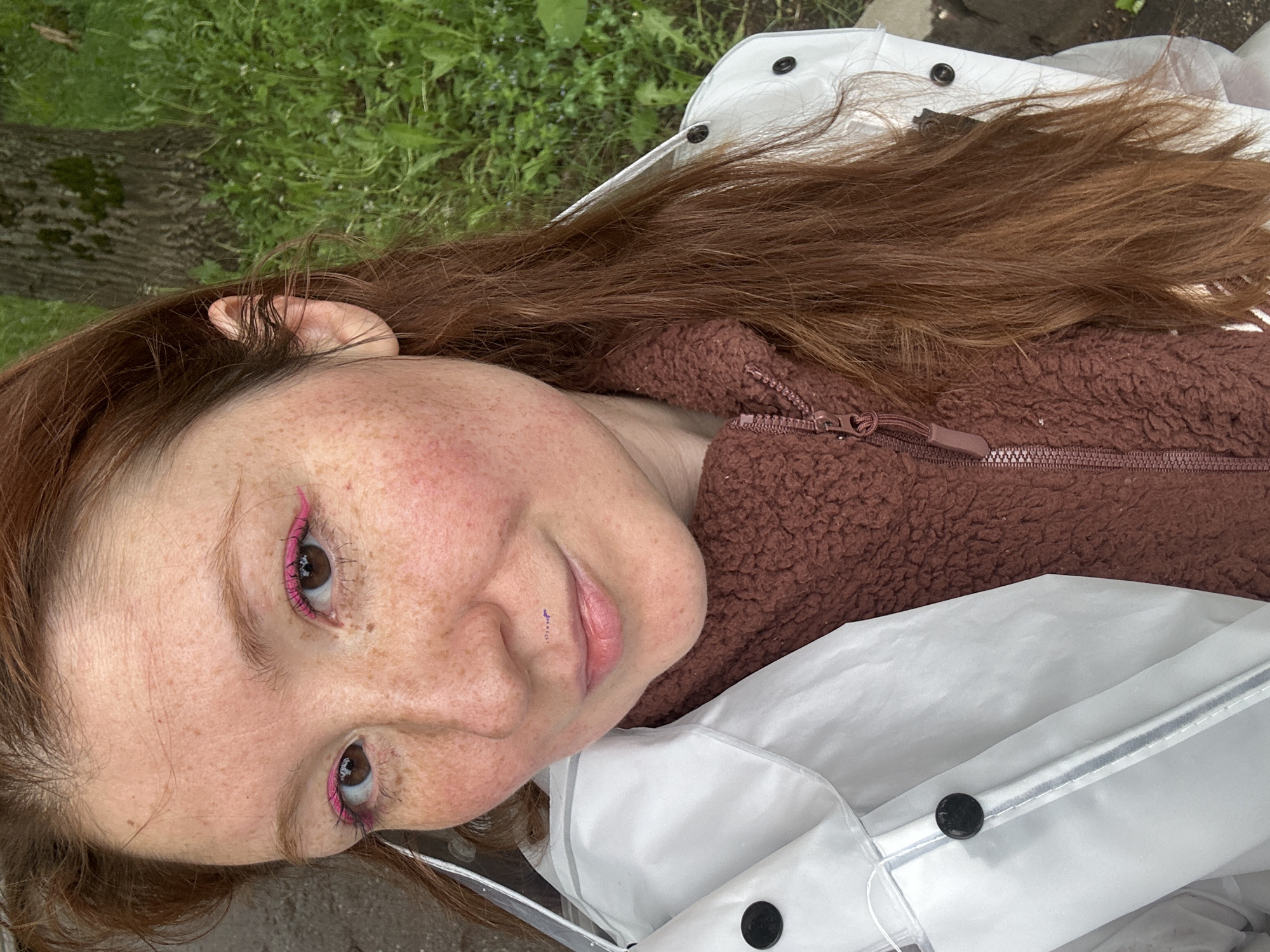 A red-haired woman with freckles and pink eyeliner is lying on the ground near a tree. She is wearing a brown fleece jacket and a white raincoat. The location is possibly in Nizhny Novgorod, Russia, but this cannot be confirmed from the image alone.