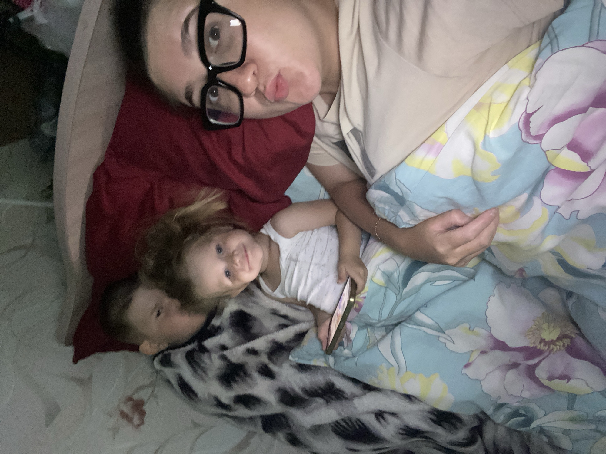 A medium shot captures a family in bed in the city of Otchor, Russia. The image is dimly lit, creating a soft, intimate atmosphere. On the right, a woman with dark hair and glasses is making a kissing face at the camera. She is wearing a light-colored t-shirt. To her left, a young child in a white tank top is smiling and holding a smartphone. The child is nestled between the woman and an older child. The older child, partially visible on the left, is wearing a patterned blanket and appears to be looking towards the phone. The family is covered in floral patterned bedding. The overall impression is one of a cozy family moment, possibly before sleep or during a relaxed time in bed.