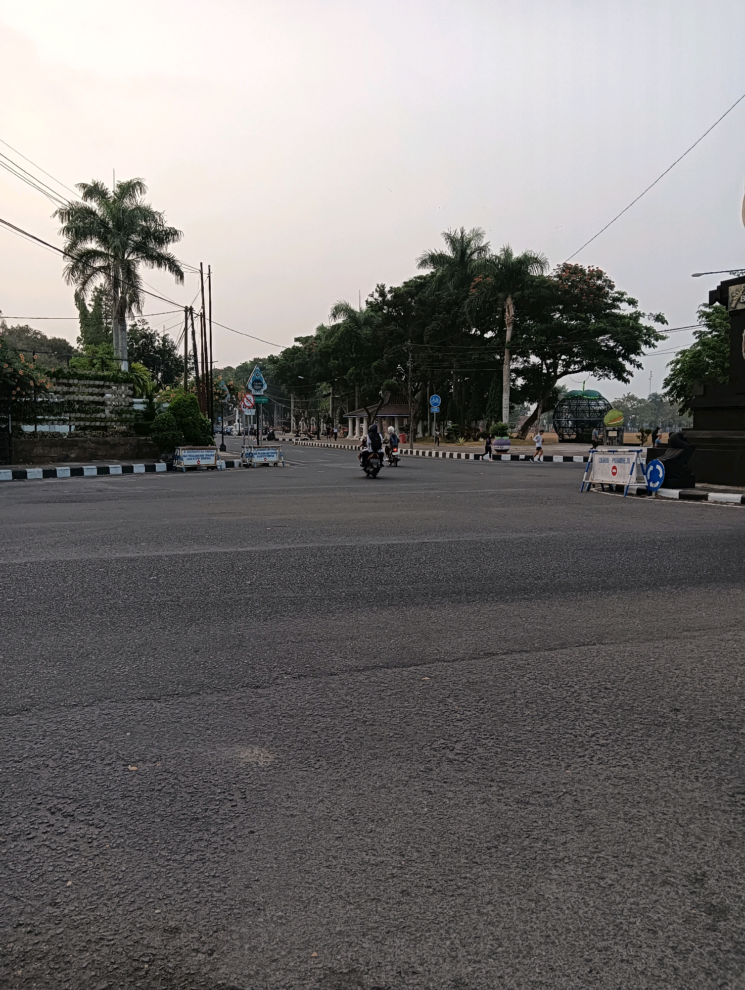 The image depicts a wide, asphalt intersection in the city of Purworejo, Indonesia, under an overcast sky. A lone motorcycle with two riders is the most prominent moving object in the frame, traveling on the road. Several other individuals are visible as small figures in the distance, some walking and others possibly running. The road is lined with a black and white striped curb, and beyond that, a raised garden bed filled with plants. Various signs are present at the intersection, including what appear to be traffic signs and temporary barriers with text on them. Trees, including palm trees, line the sides of the road, and utility poles with visible wires are scattered throughout the scene. A large, spherical sculpture or art installation is visible in the background on the right side of the road. The overall impression is that of a quiet urban intersection, possibly in the early morning or late afternoon, with minimal activity.