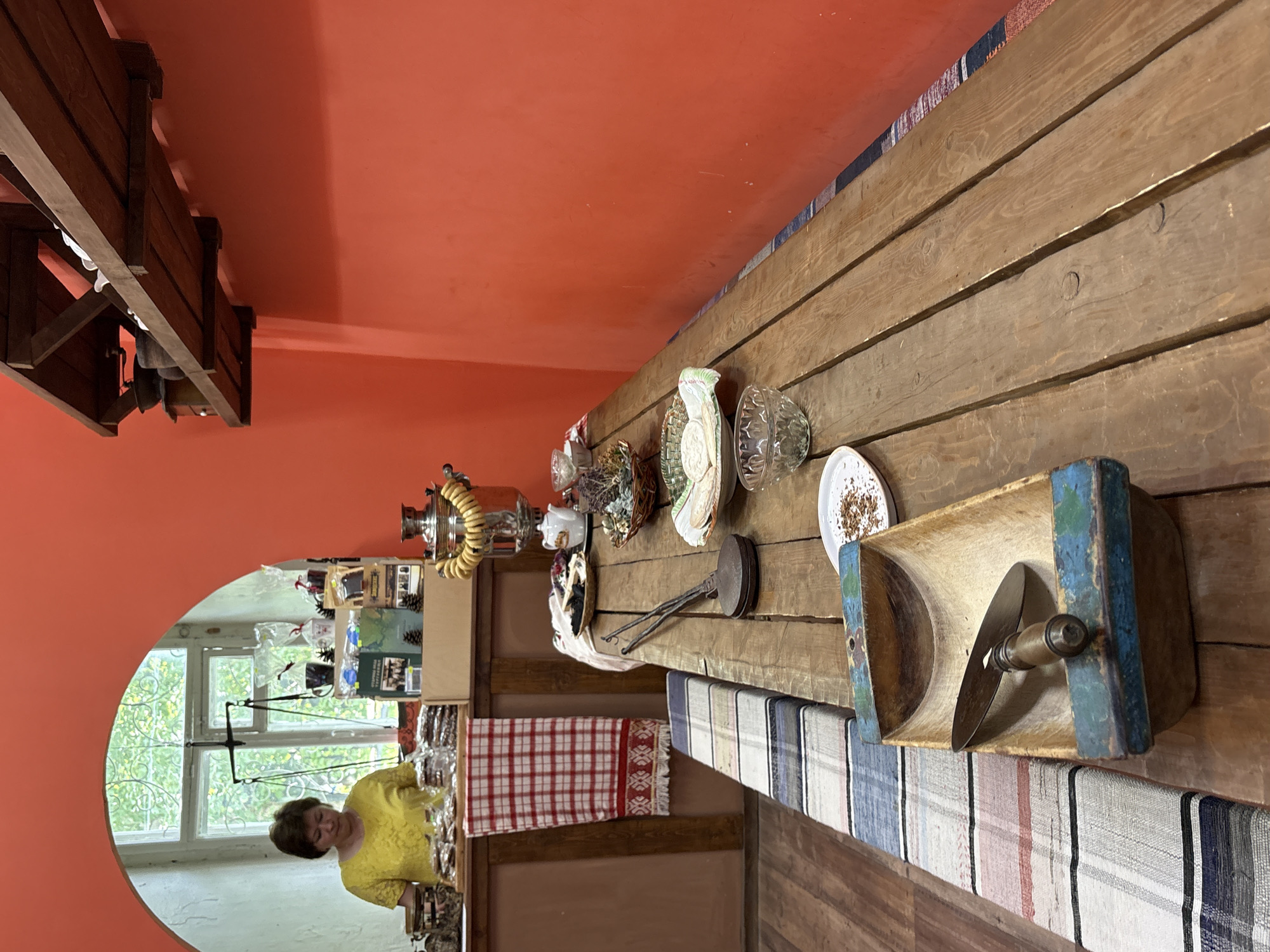 The image depicts the interior of a rustic room with a prominent orange wall and a wooden table laid out with various items. In the background, visible through an arched opening, a woman in a yellow shirt stands behind a counter. The counter displays goods, and a window with green foliage can be seen behind her. The foreground features a long, rough-hewn wooden table covered with a colorful striped runner. On the table are several glass bowls, a samovar adorned with a braided ring of dough, a small white teapot, a basket of dried herbs, a vintage waffle iron, and a wooden trough containing a knife. The environment suggests a traditional Russian setting, possibly a shop or a domestic space in Suksun, Russia, known for its local crafts and historical samovar-making tradition.