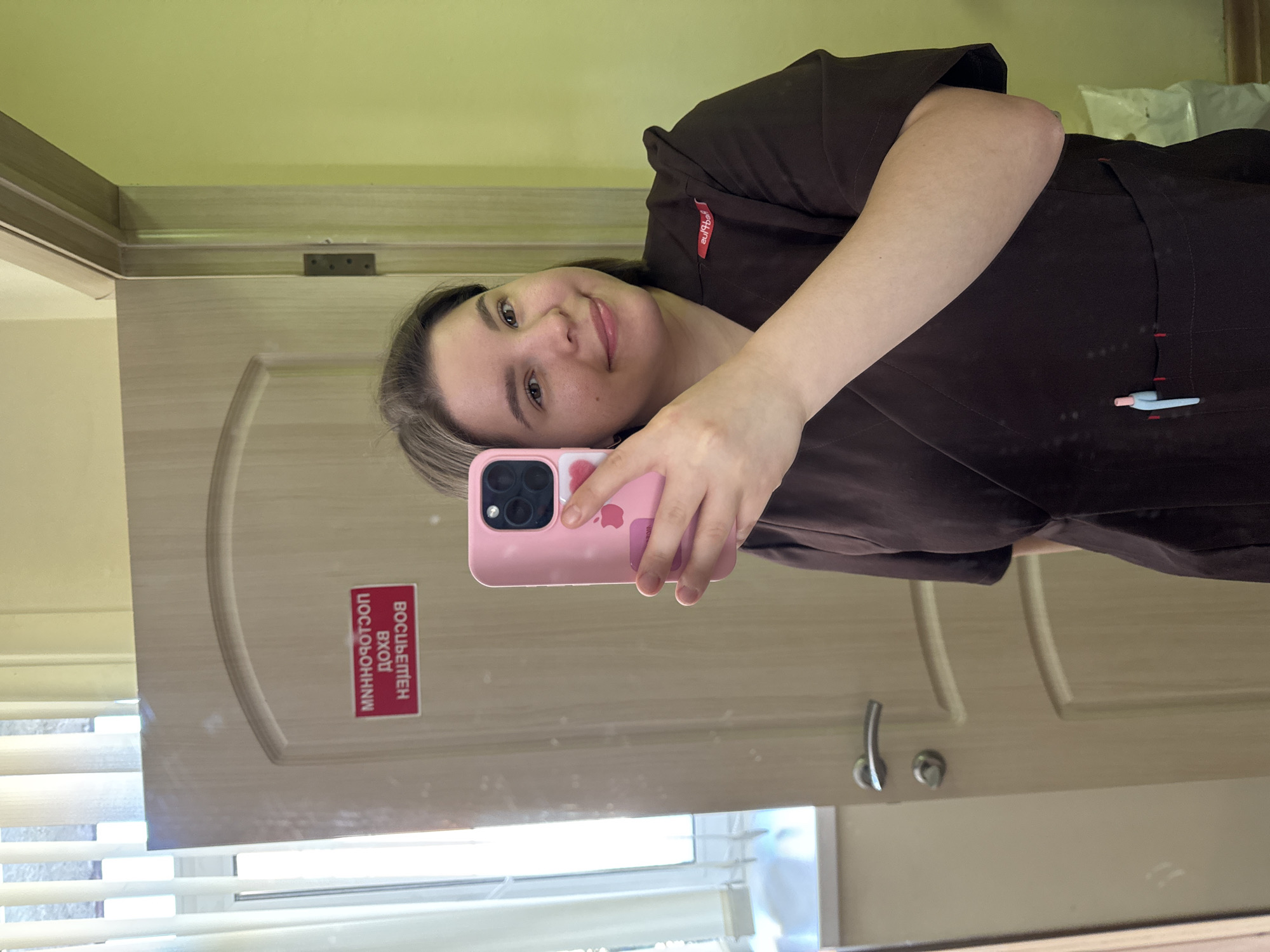 A woman is taking a selfie in front of a mirror, holding a pink iPhone. She is wearing a brown scrub top. In the mirror's reflection, a door is visible with a sign in Russian that reads "ВОСВЕЩЕН ВХОД ПОСТОРОННИМ." The background has a beige-colored wall. Based on the writing on the door and the overall context, the location is likely Kazan, Russia.