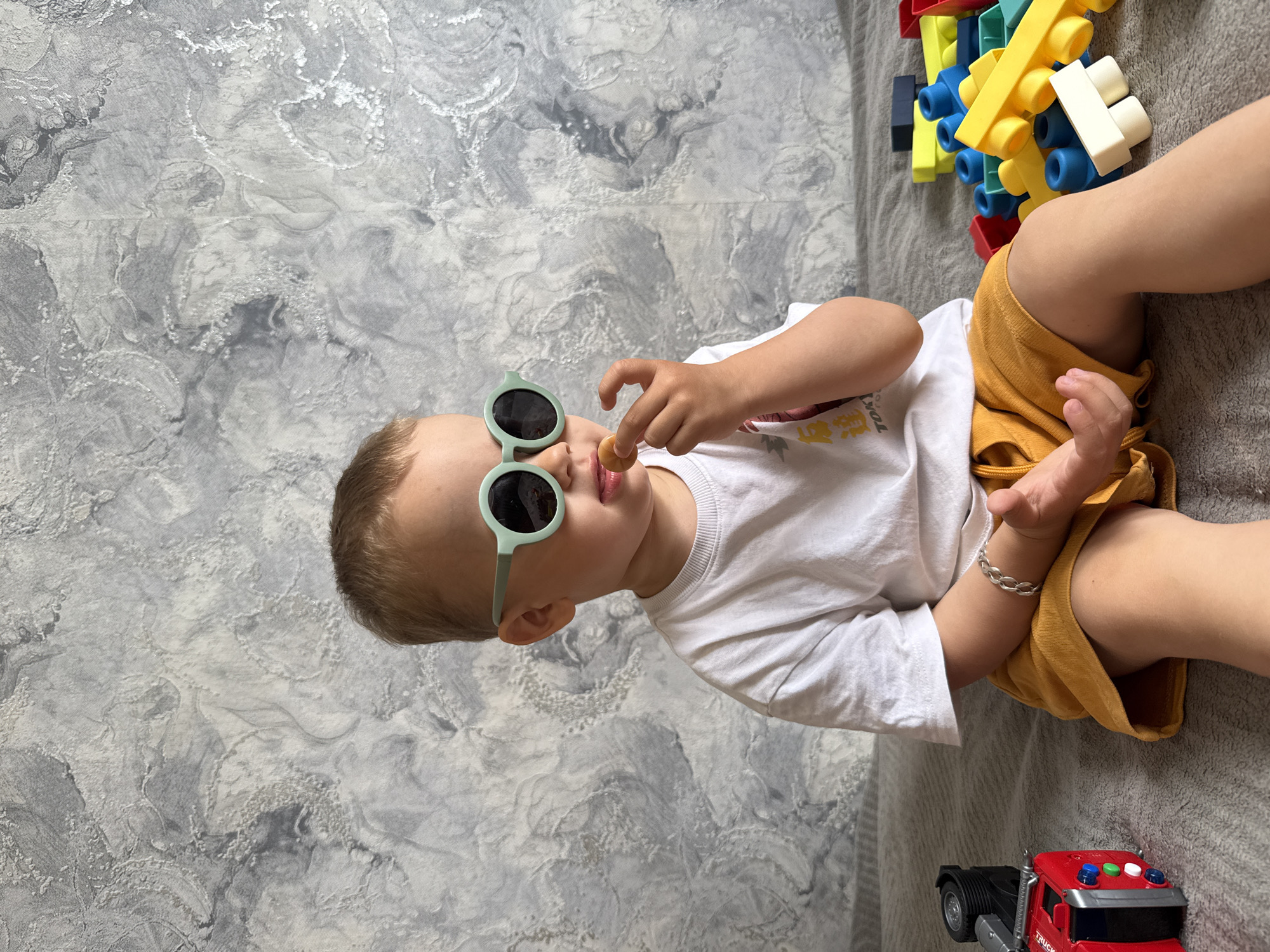 A child with fair skin and brown hair is lying on a gray textured surface. The child is wearing round, light green sunglasses, a white t-shirt with yellow lettering, and mustard-colored shorts. The child is holding a small, light brown object in their right hand, near their mouth. Beside the child are colorful plastic building blocks and a toy red fire engine truck. The background features a gray and white marbled wall texture. There is no evidence to support the claim that the image was taken in the city of Rusanovo, Russia.