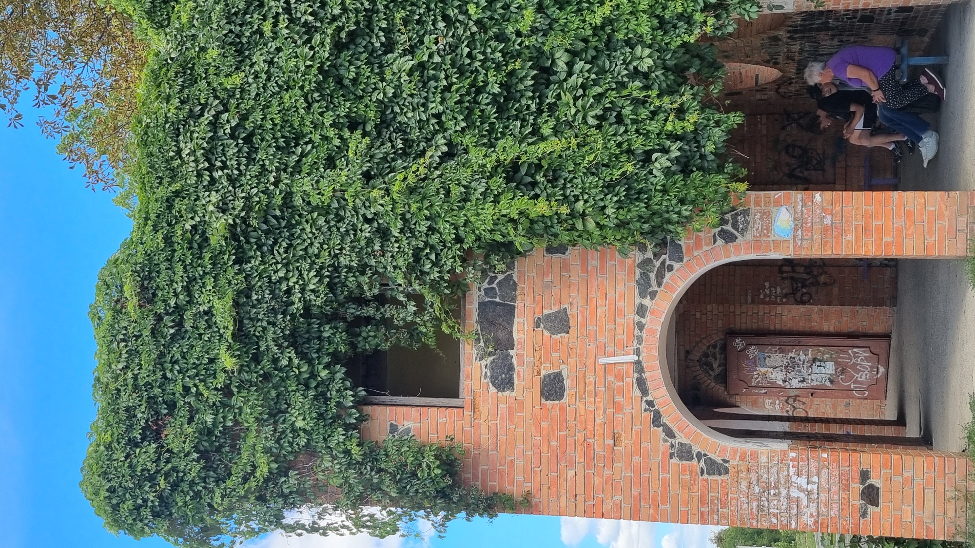 In the city of Smila, Ukraine, a brick building, heavily overgrown with green ivy, stands under a blue sky with scattered clouds. The building has an arched entranceway with graffiti-covered doors and walls. Inside the arched structure, three individuals are seated on a bench. Two younger individuals, one male and one female, sit to the left, both looking at their phones. To the right, an older woman in a purple shirt and patterned pants sits looking forward. The building appears to be abandoned, with a vacant window opening visible on the upper level, surrounded by more ivy. The overall scene suggests an outdoor, slightly dilapidated public space.