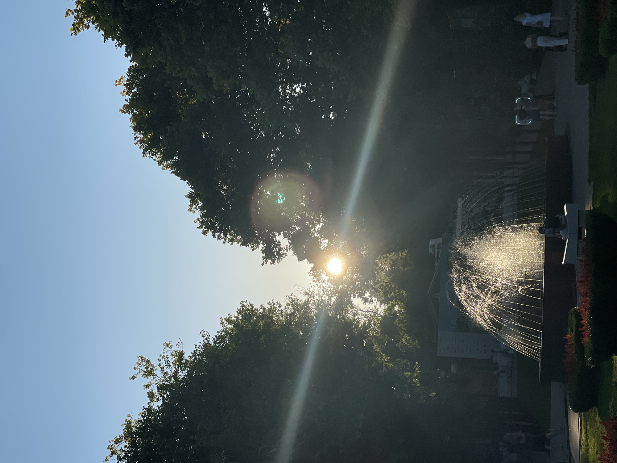 The image depicts a fountain in a park setting, likely in Sochi, Russia, given the context provided. The sun is setting or rising, casting a warm glow and creating lens flare across the scene, particularly through the trees that frame the sky. The fountain is the central subject, with water spraying upwards and catching the sunlight, creating a shimmering effect. Several people are visible in the background, some walking and others standing, suggesting a public space. The environment appears to be a well-maintained park with lush greenery and manicured bushes. The overall mood is serene and tranquil, with the natural light playing a significant role in the composition.