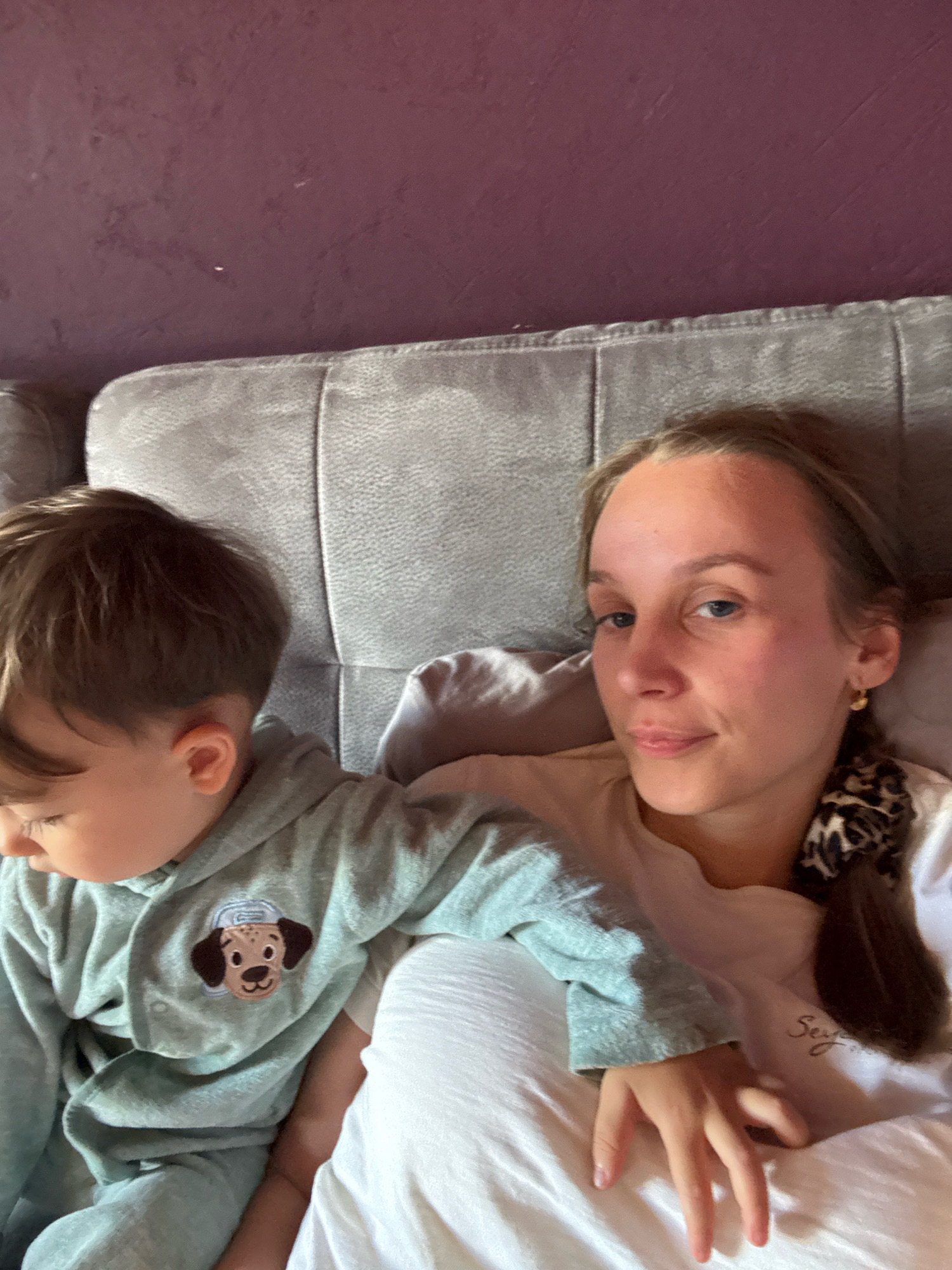 The media file features a young child and an adult woman lying down on a grey couch or bed. The child, facing away from the camera and towards the left, is wearing a light blue hooded tracksuit with a cartoon dog face embroidered on the chest. The adult woman is positioned to the right of the child, looking towards the camera with a slight smile. She has light skin, blue eyes, and her hair is pulled back into a braid with a leopard-print scrunchie. She is wearing a light-colored t-shirt. The background consists of a dark purple wall. The scene appears to be indoors, possibly in a living room or bedroom. The lighting suggests it could be daytime. The overall mood seems relaxed and intimate, with the two subjects comfortably close. There is some faint text on the woman's t-shirt, but it is not clearly legible. The location is identified as Überherrn, Germany.