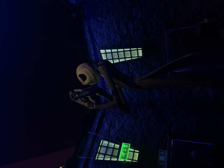 Jack Skellington, the Pumpkin King, holds something in his hands. He's rendered in a dark, animated style, set against a building with textured walls in shades of blue. Square windows with bright green lights punctuate the scene, including one with an illuminated emergency exit symbol. A portion of the image is immersed in deep shadow, adding to the eerie atmosphere.