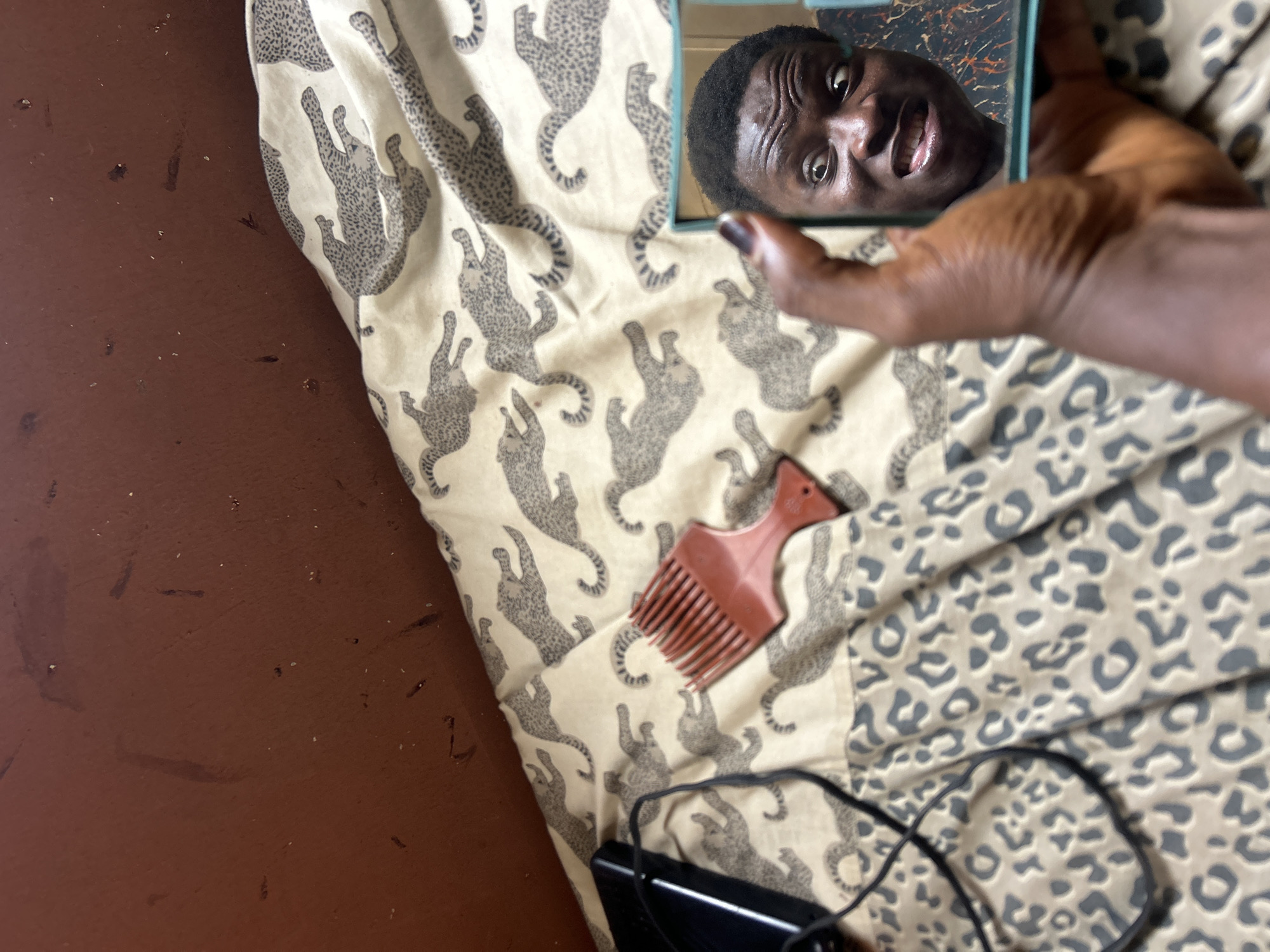 An overhead, angled shot captures a person's reflection in a small handheld mirror, placed on a patterned fabric. The reflected face, belonging to a dark-skinned individual with short, dark, textured hair, shows wide eyes, a wrinkled forehead, and a slightly open mouth revealing teeth, suggesting an expressive or surprised look. A hand, also dark-skinned, holds the mirror's teal-colored frame, with a hint of dark nail polish visible on one finger.
The fabric beneath the mirror features two distinct animal print patterns: one with stylized, crawling leopard figures on a light background, and another with more traditional, abstract leopard spots. A reddish-brown afro pick-style comb rests on the patterned fabric near the center. Below it, a black electronic device, possibly a power bank or phone, with a coiled black cable, is partially visible. The background to the left consists of a dark reddish-brown surface, resembling a floor, which appears somewhat worn with scattered specks and smudges. The overall scene suggests an intimate, casual, indoor setting. There is no visible text in the image.