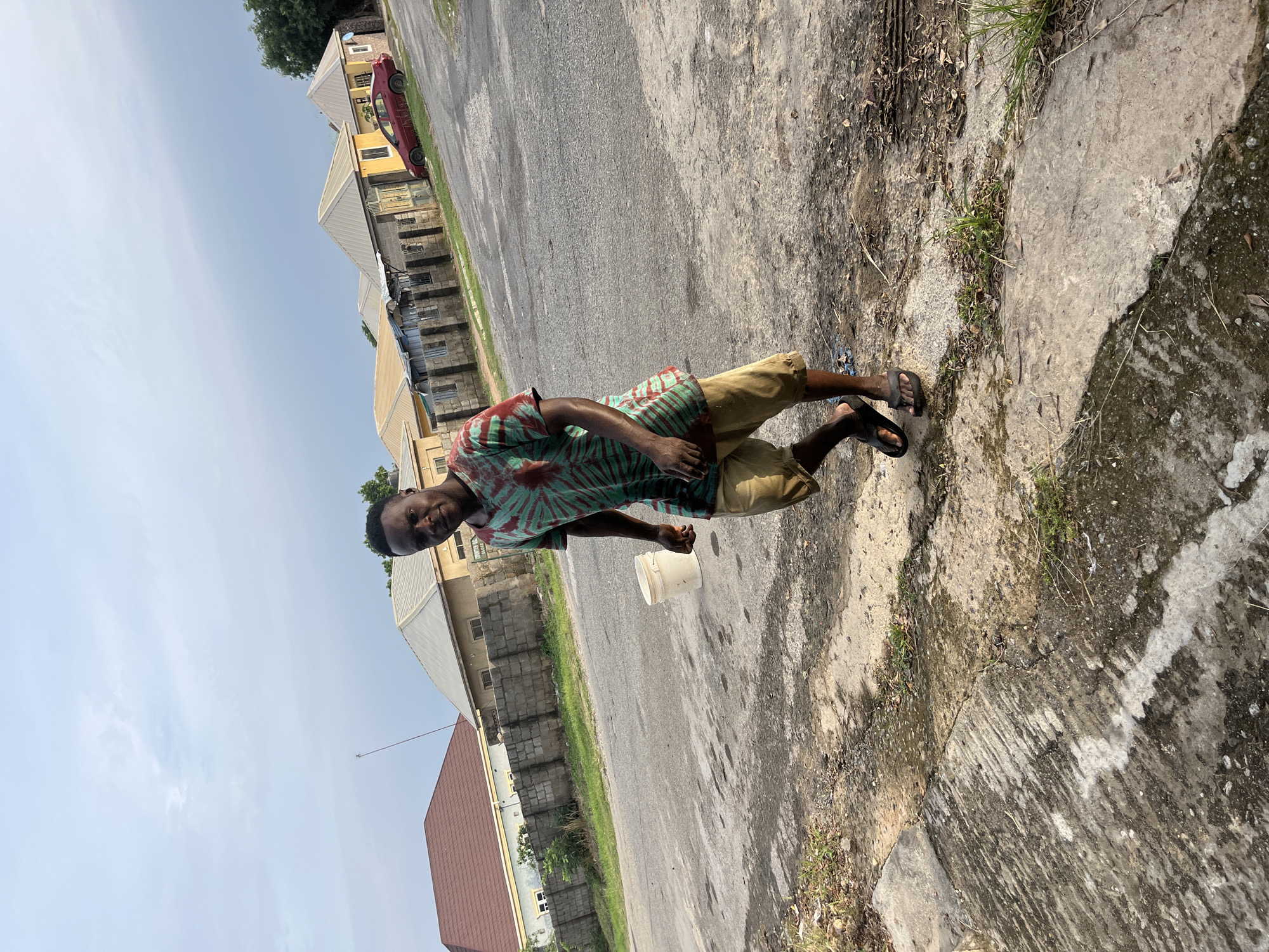 A young man walks on a dirty, paved road in front of residential buildings in the city of Abuja, Nigeria. The man is wearing a green and red tie-dye t-shirt and light brown shorts. He is walking towards the viewer, with his head turned slightly to his left. In the background, several houses with different colored roofs and walls are visible. A red car is parked on the right side of the road, in front of one of the houses. A white bucket is on the ground to the left of the man. The sky is overcast with clouds.