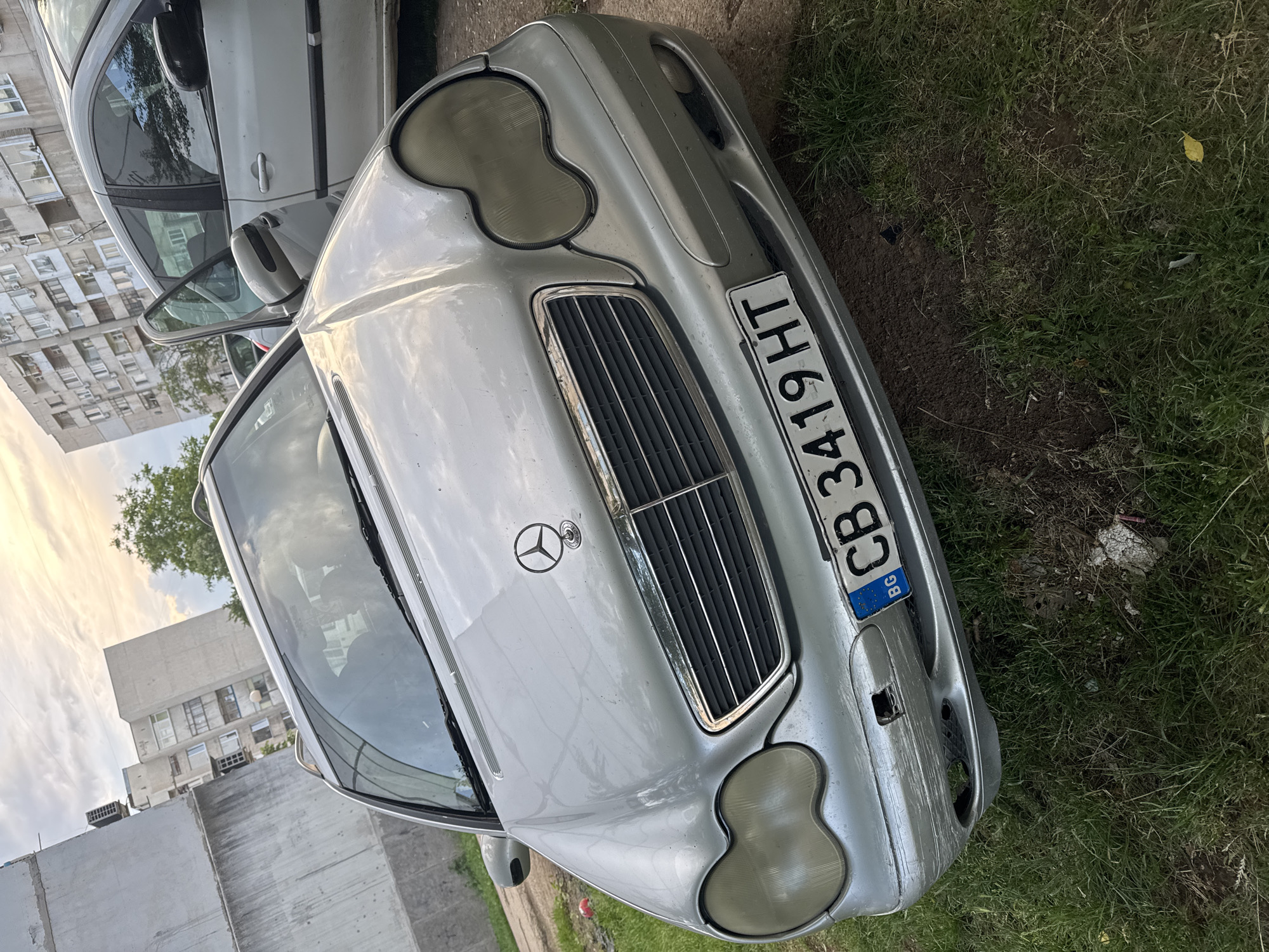A silver Mercedes-Benz car, likely a C-Class (W203 generation) or CLK-Class, is the central subject of this image, positioned diagonally. It appears parked on a patch of grass and dirt next to a paved area. The car's front features the iconic Mercedes-Benz star emblem on the grille, which is metallic and has vertical slats. Its headlights are noticeably fogged or yellowed, indicating wear and tear. The front bumper shows visible scuffs and some damage on the lower right side.
The license plate, "CB 3419 HT," is prominently displayed on the front bumper. The "CB" designation, along with the "BG" country code within the blue EU strip, confirms the vehicle is registered in Sofia, Bulgaria.
In the background, several multi-story residential buildings are visible, characterized by numerous windows and a somewhat utilitarian architectural style common in urban settings, aligning with the Sofia, Bulgaria context. Another light-colored car is partially visible behind the Mercedes. The sky is partly cloudy, suggesting it's daytime, possibly late afternoon, given the diffused light. No people or specific activities are depicted, giving the scene a quiet, almost neglected appearance, particularly due to the car's condition and its parking location off a clear road.