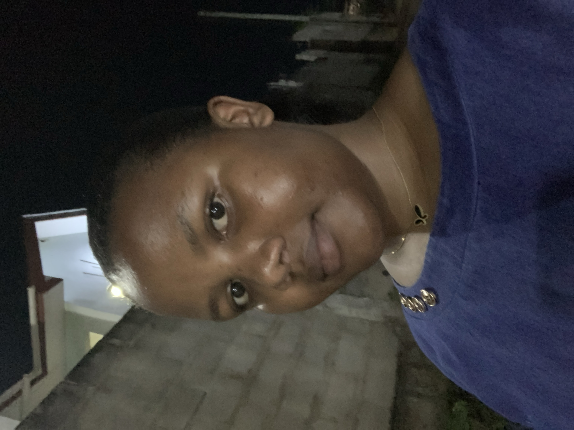 A person is positioned in front of a building, presumably in the city of Dar es-Salaam, Tanzania. It is nighttime, with dark skies visible in the upper portion of the frame. The person, likely an adult, is facing the camera with their head tilted slightly. Their facial features, including eyes, nose, and mouth, are visible. They are wearing a garment with decorative buttons near the neck. A building with lights and a balcony-like structure can be seen in the background. The ground appears to be made of stone or concrete.