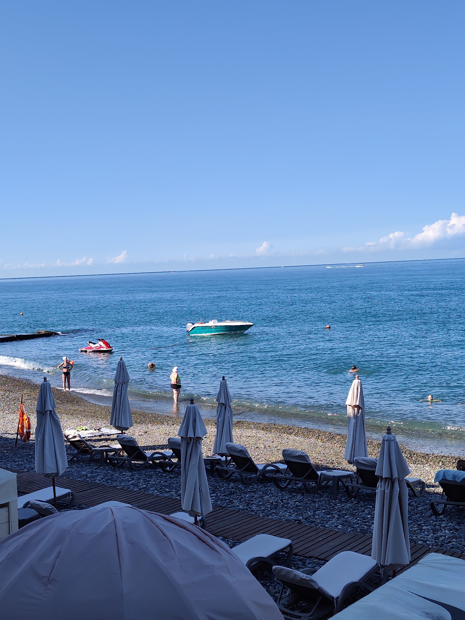 A wide shot captures a clear, blue sky over the Black Sea in Sochi, Russia. The foreground is dominated by a paved area with several closed beach umbrellas and empty lounge chairs. A large, pink beach umbrella is prominently featured in the lower left. Beyond this, a pebble beach is visible, with more closed umbrellas and lounge chairs.
The sea is calm and a bright blue, with gentle waves lapping at the shore. A few people are in the water. Near the shore, a red jet ski is partially submerged, and a turquoise motorboat is anchored further out. Two people are visible standing in the shallow water on the beach. In the distance, several more boats are present on the sea. The horizon is clear and the weather appears to be sunny.
