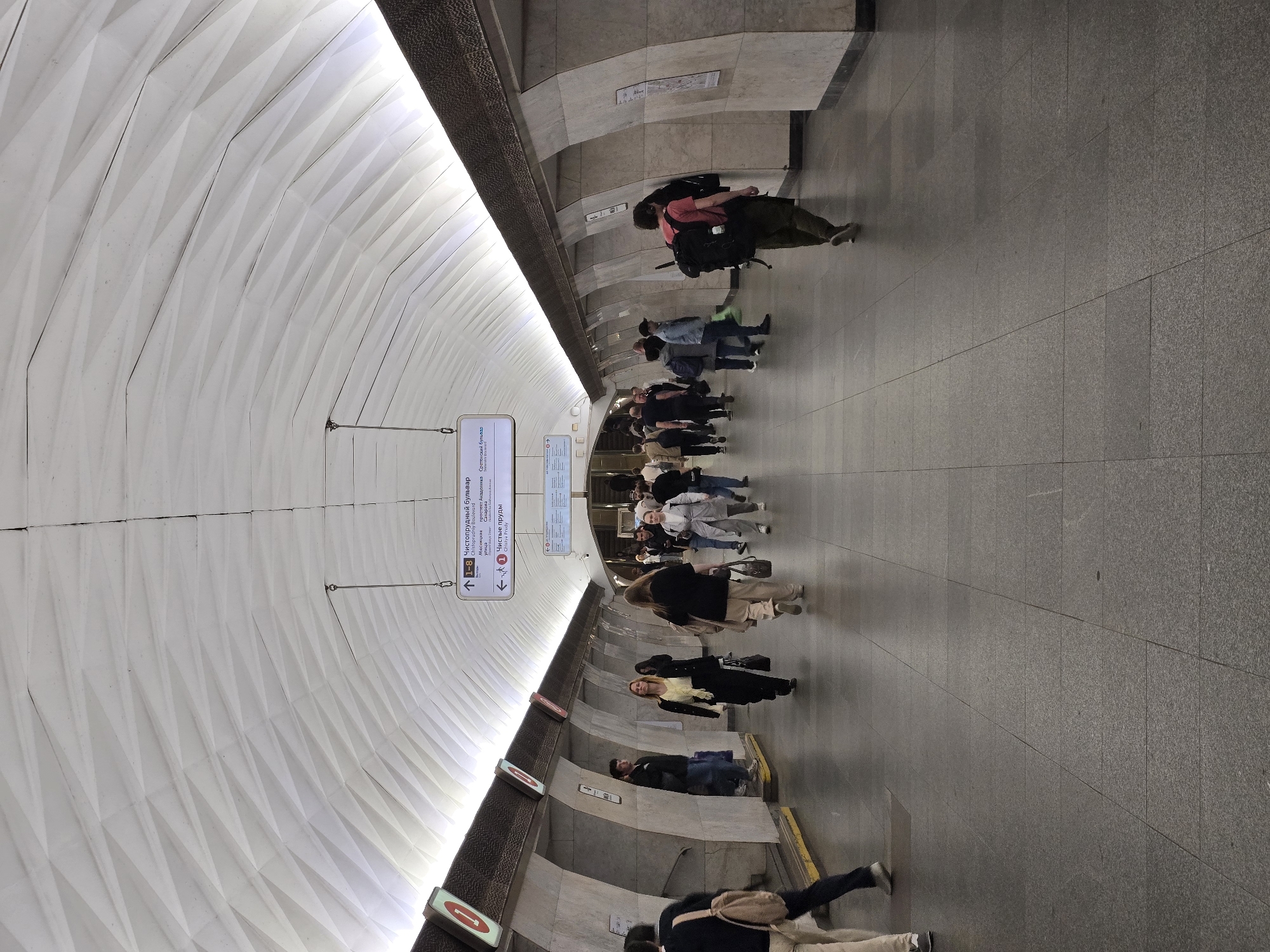The image shows an underground pedestrian walkway with numerous people traversing it. The architectural design includes a distinctive, faceted, white ceiling that curves downward towards the sides. A sign indicates directions to "Chistoprudniy Boulevard," "Myasnitskaya," and other locations in Moscow, Russia, specifying "Чистые пруды" (Chistye Prudy) as well. The walkway is paved with gray stone or concrete. People are walking in both directions along the passageway.