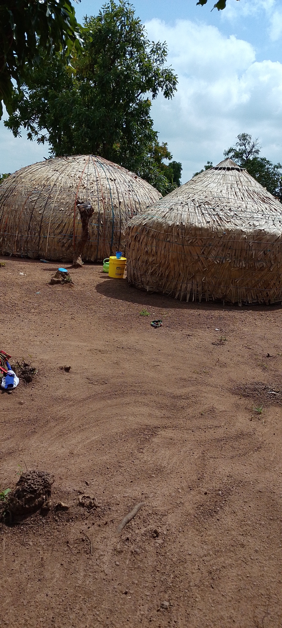 Two domed, thatched-roof structures, resembling huts, are situated on reddish-brown, sandy ground. The structure on the left is larger and wider, with vertical poles supporting its thatched walls, which are made from palm fronds. A smaller, similar structure is located to the right and slightly behind the first. A rough wooden post stands between the two huts. In front of the left hut, a small blue object rests on a rock formation. To the right of the larger hut, a yellow bucket with a blue lid and a smaller green container are placed on the ground. Scattered on the ground are a few items of clothing, including a white garment with blue and red accents, and some debris. In the background, lush green trees with full foliage form a canopy against a partly cloudy sky. The scene appears to be an outdoor settlement, possibly in a rural or semi-rural area.