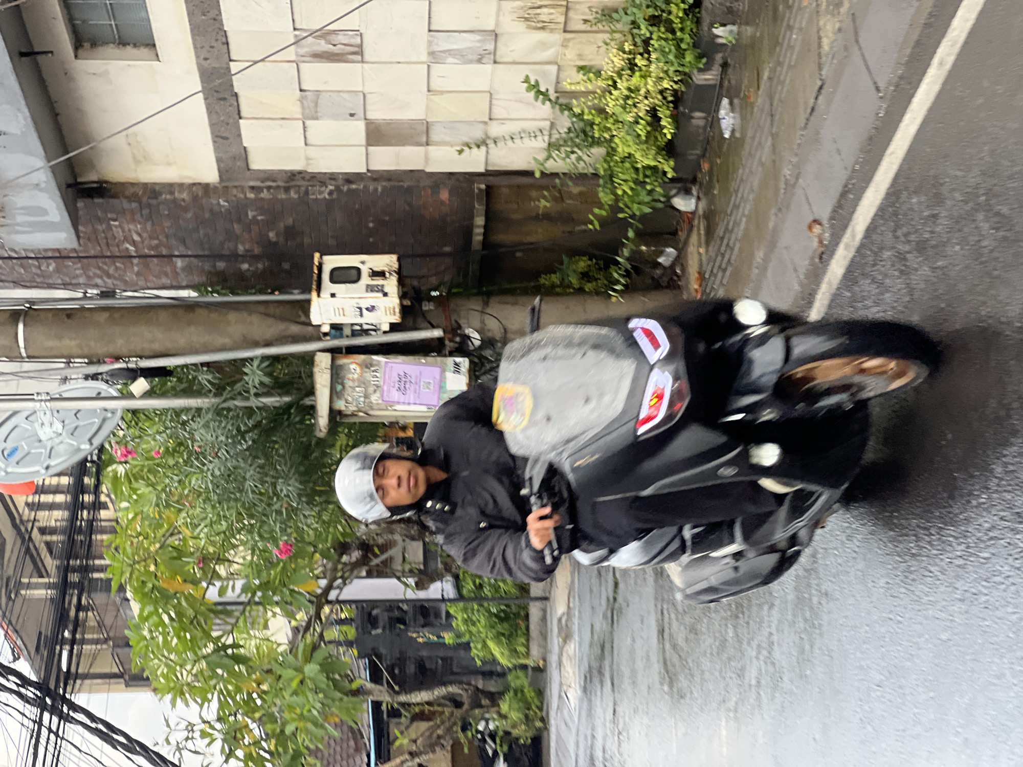 A person wearing a silver helmet and a black jacket is riding a black scooter on a wet street in North Kuta, Indonesia. The scooter has red taillights and a clear windshield. The person's face is visible, and they are looking forward. The street is asphalt and appears to be in a city, with buildings and trees in the background. There are electrical wires overhead and a utility box on the side of a building. The overall lighting suggests it is daytime, possibly overcast or after rain.