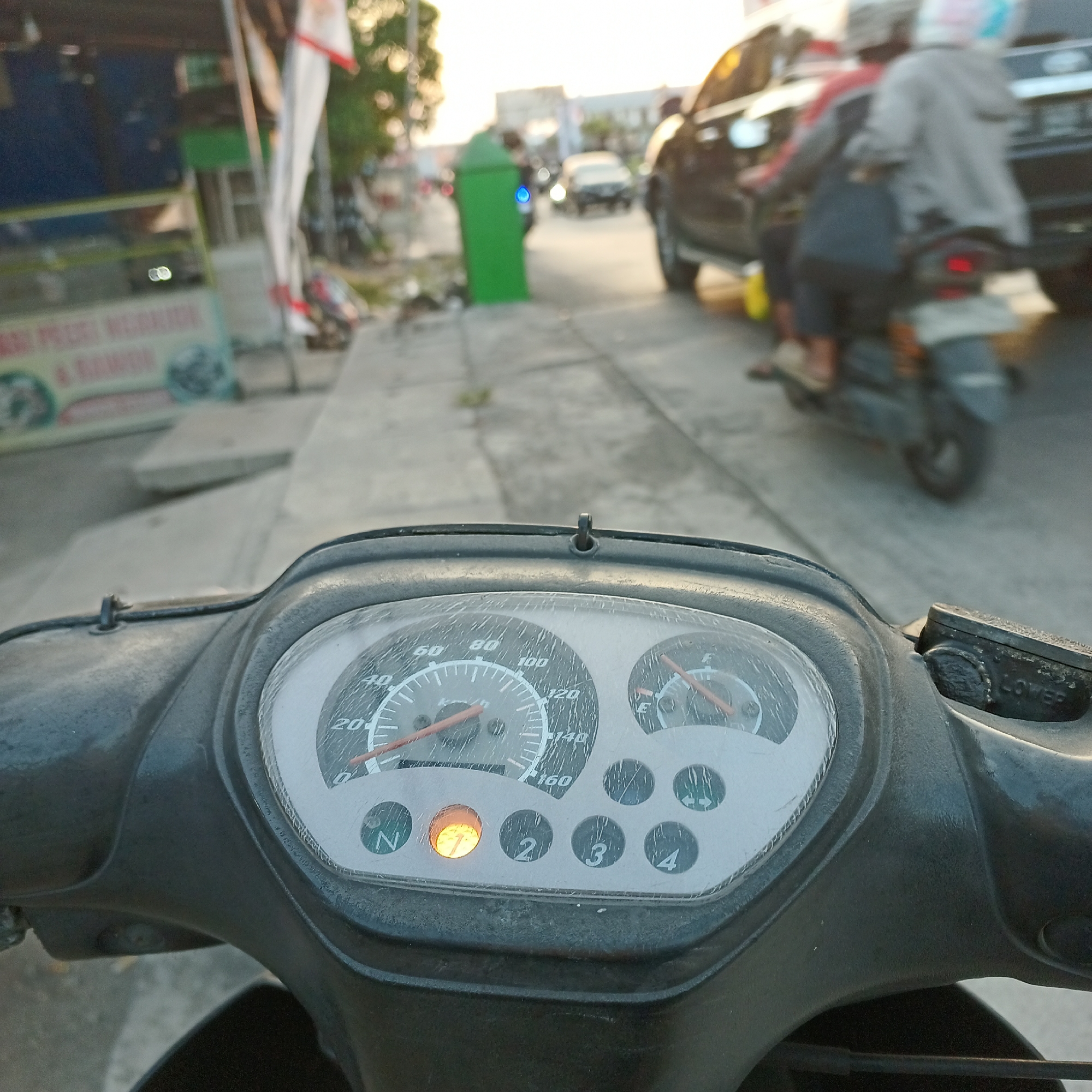 The image shows a close-up view of a motorcycle's dashboard, with a speedometer indicating a speed between 0 and 20 km/h. The neutral indicator light is illuminated orange. In the background, traffic can be seen on a street in what appears to be an urban or suburban area. A green trash bin is visible on the left side of the road, and two people on a motorcycle are in motion. The setting is consistent with Kepuh Kiriman, Indonesia, given the context of the request. The overall scene depicts a moment of slowed traffic or a brief stop, possibly at an intersection or during congestion. The time of day appears to be late afternoon or early evening, judging by the warm lighting and shadows.