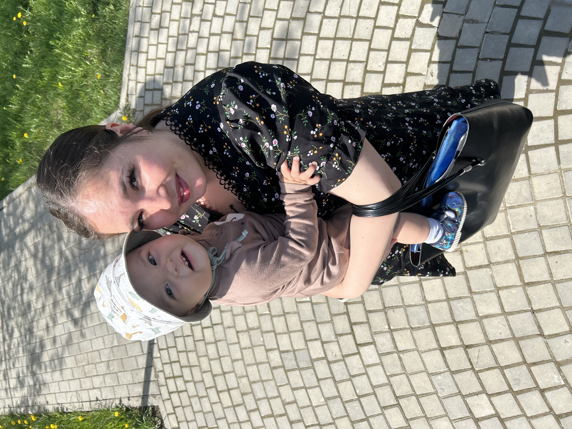 A woman is holding a baby in an outdoor setting. The woman is wearing a black floral dress and has her hair pulled back. The baby is wearing a light brown long-sleeved shirt and a patterned hat. They are both smiling at the camera. The background consists of a paved area with grass and dandelions visible in the periphery. There is a black bag on the ground next to the woman's legs. The location appears to be a park or public area within the city of Votkinsk, Russia.