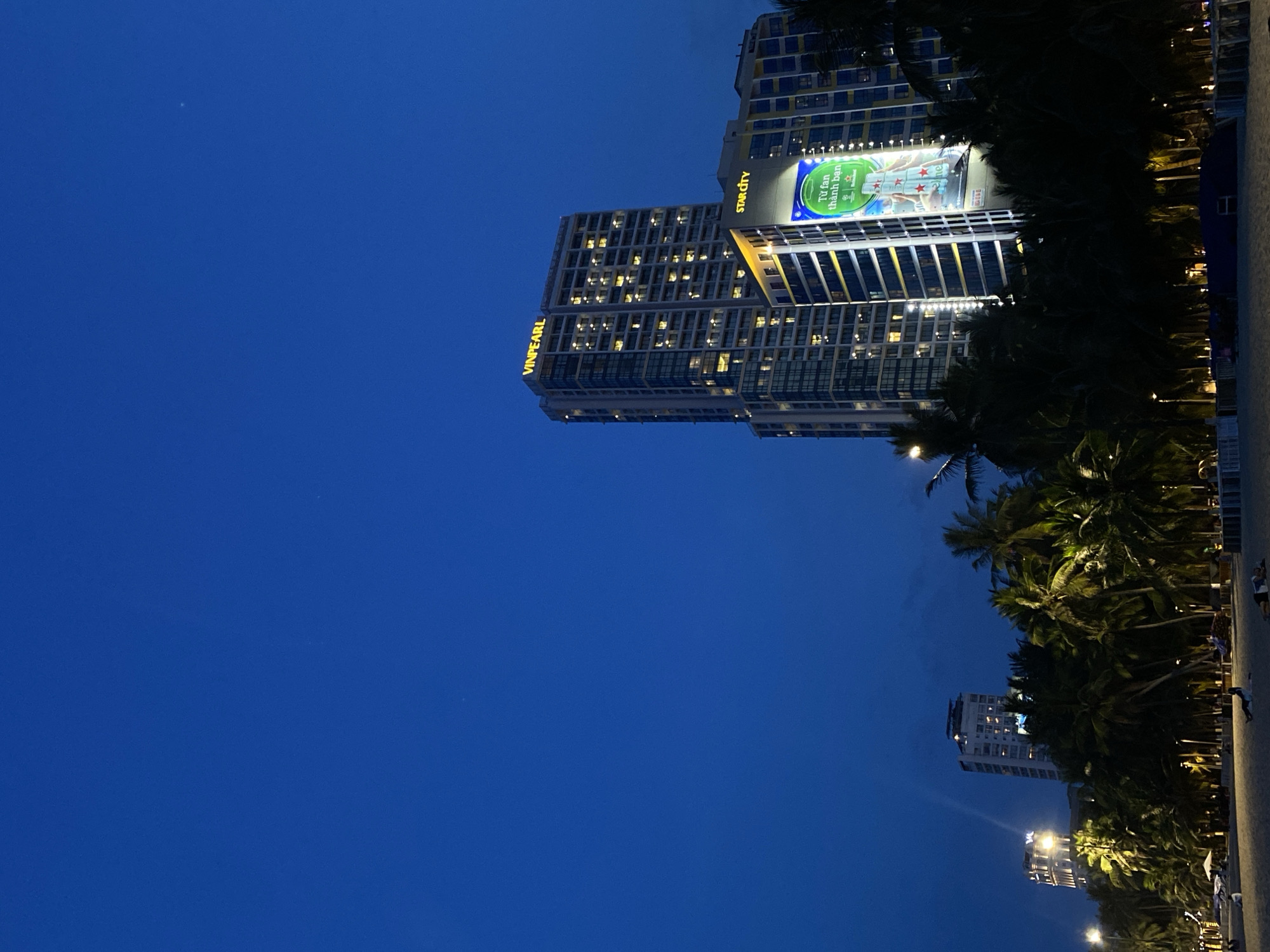 This media file depicts a twilight scene in Nha Trang Ward, Vietnam, likely a beachfront area. The sky is a deep, dark blue, suggesting the transition from day to night.
The primary subjects are a series of modern high-rise buildings illuminated by many lit windows. One prominent building displays a large, brightly lit billboard advertising a product, with Vietnamese text visible. Another building has "STAR CITY" written on its facade. The word "VINPEARL" is also visible in large yellow lettering on another building.
Lush palm trees line the foreground, their dark silhouettes contrasting with the lit buildings. The presence of palm trees and the general ambiance suggest a tropical or subtropical coastal location.
The scene is serene, with no overt activity depicted other than the static presence of the buildings and vegetation. The overall mood is calm and perhaps a little mysterious due to the low light conditions. There are subtle hints of people in the distance, possibly walking along a beach or promenade, but they are not clearly discernible. Streetlights are also visible, casting a warm glow. The time of day is clearly evening or dusk.