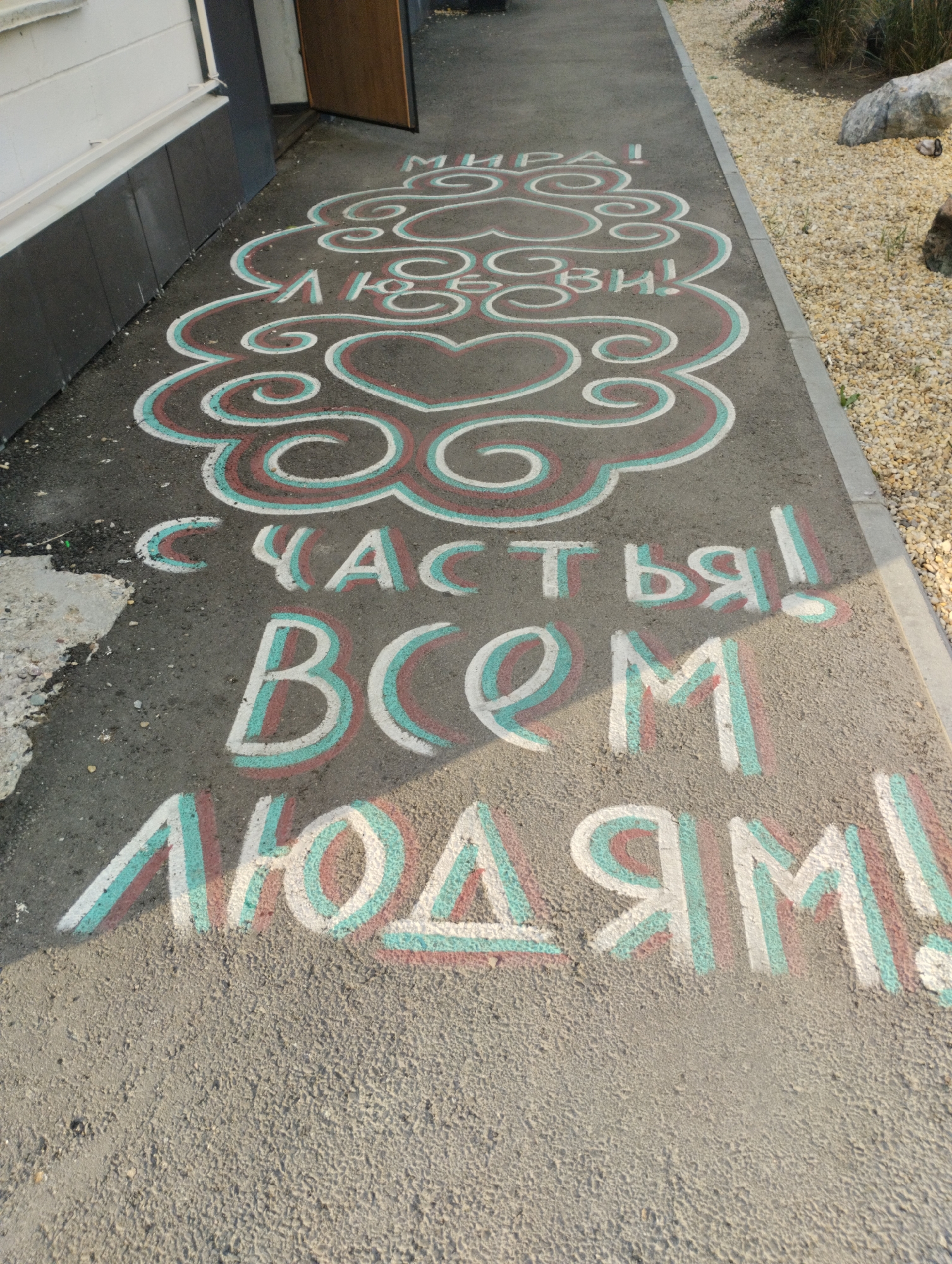 The image depicts a chalk drawing on an asphalt surface, likely a sidewalk or pathway, with a building on the left and gravel and vegetation on the right. The chalk drawing is in three lines. The first line reads "МИРА!" in white chalk with red and teal outlines, above a decorative cloud-like design with a heart in the center. The second line reads "ЛЮБВИ!" in white chalk with red and teal outlines, also above the decorative design. The third line, larger and more prominent, reads "СЧАСТЬЯ ВСЕМ ЛЮДЯМ!" in white chalk with red and teal outlines. The building on the left is white with dark gray lower panels and a dark brown door that is ajar. The right side of the image shows light-colored gravel, sparse green vegetation, and some rocks, including a large boulder. The sun is casting shadows, indicating it is likely daytime. The scene appears to be in an outdoor public space, possibly near an entrance to a building or in a recreational area. The chalk drawing is a message of peace, love, and happiness. There are no people visible in the image.