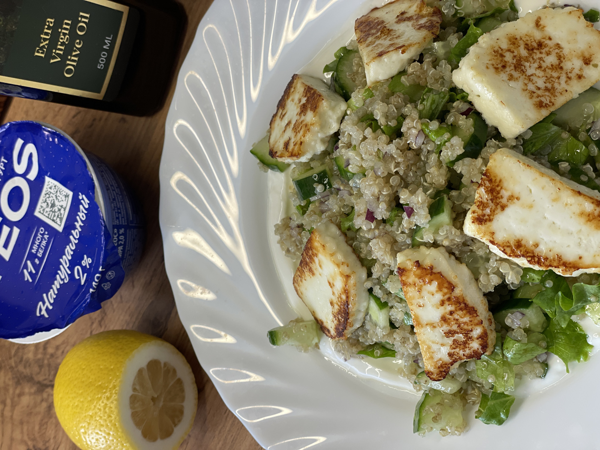 The image displays a meal arranged on a light brown wooden surface, located in Chaikovsky, Russia. In the foreground, a white ceramic plate contains a salad primarily composed of quinoa, chopped cucumber, green lettuce, small pieces of red onion, and multiple pan-fried or grilled halloumi cheese cubes exhibiting distinct golden-brown grill marks. A creamy white dressing is visible mixed with the salad ingredients. In the upper left background, a yellow lemon, cut in half horizontally, reveals its internal structure. Adjacent to the lemon and partially obscured, a blue and white plastic container of "EOS" brand natural yogurt, bearing Russian text "ЙОГУРТ," "11 Г МНОГО БЕЛКА" (11g high protein), and "Натуральный 2%," is visible. To the far right, a dark glass bottle with a label indicating "Extra Virgin Olive Oil" and "500 ML" is present. No individuals are depicted.