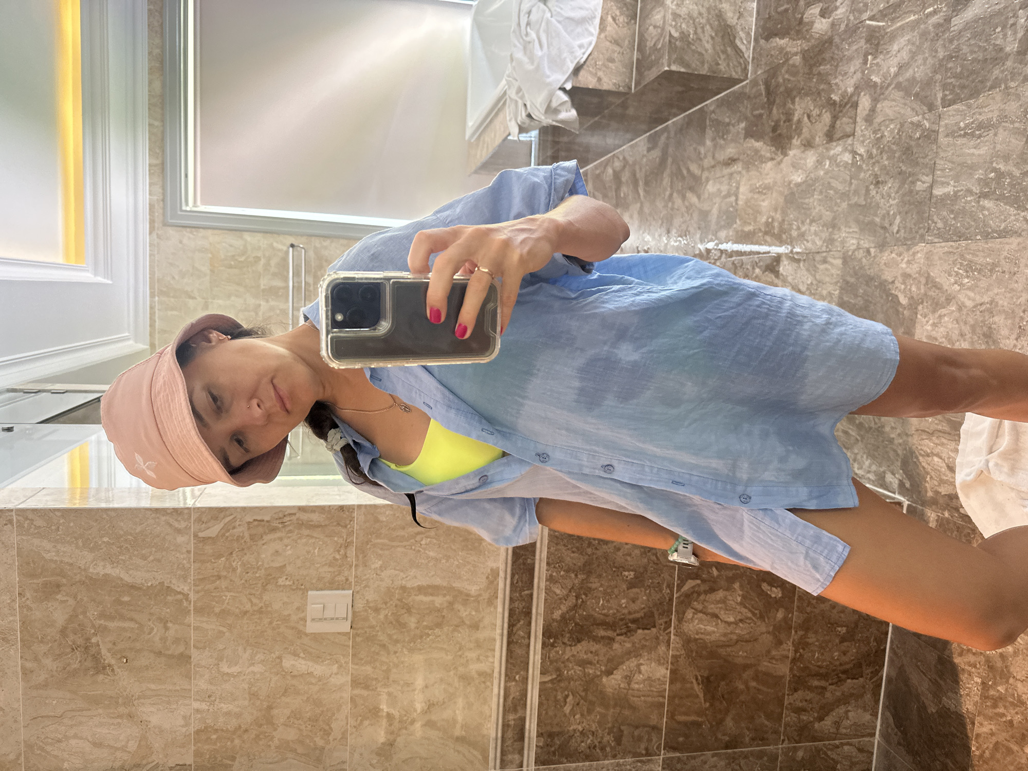 In Nha Trang Ward, Vietnam, a forensic image captures a female adult taking a mirror selfie in a bathroom. The subject, positioned centrally, wears a light pink bucket hat with a white logo, a neon yellow top, and an unbuttoned oversized light blue linen shirt. She displays a small nose piercing, a gold-colored necklace with a pendant, a white smartwatch with a teal band on her left wrist, and a gold ring on her right ring finger. Her right hand, with magenta-painted fingernails, holds a smartphone in a clear case, reflecting her face. The bathroom features light brown marble-patterned wall and floor tiles, a decorative darker brown border tile, and white crown molding with integrated warm-toned lighting visible in the ceiling. A white light switch panel is present on the left wall. In the reflection, a metal towel rack and a white bathtub with tiled steps are visible on the right. A white towel is partially visible on the tiled floor near the subject's feet.