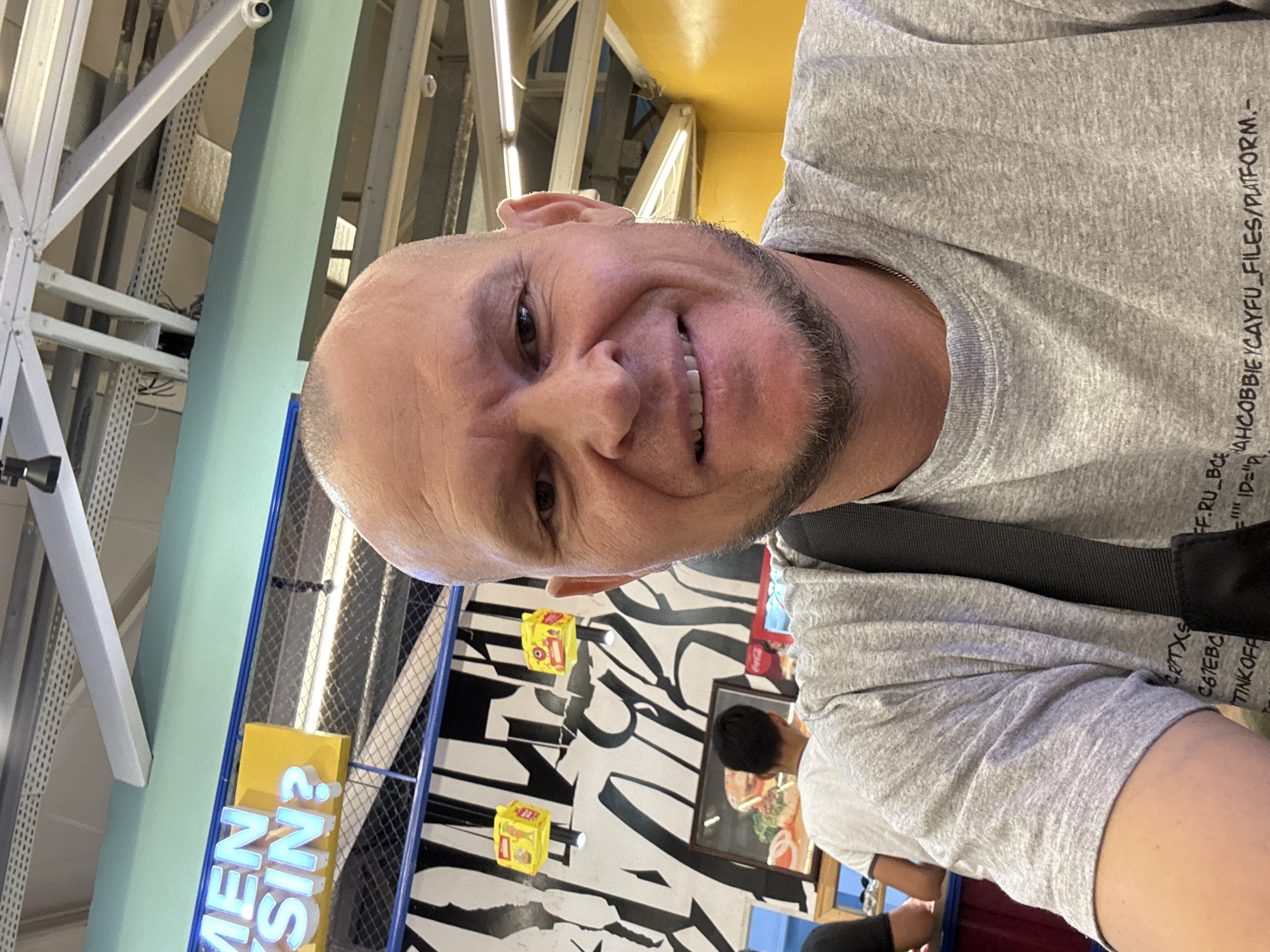 A man with a shaved head and a goatee is taking a selfie in an indoor public space, likely a food court or restaurant. He is wearing a grey t-shirt with text printed on it. The background features a bright yellow wall, a black and white striped wall mural, and signage. One sign, illuminated with blue neon lights, appears to read "MEN SIN?". There are also yellow boxes with images of food products hanging from the ceiling. The ceiling structure with exposed metal beams and lights is visible. In the background, other people are present, including someone wearing a white t-shirt facing away from the camera, and a glimpse of a menu or advertisement featuring a burger. The lighting is bright, suggesting an indoor commercial area. The location is identified as Oskemen, Kazakhstan.