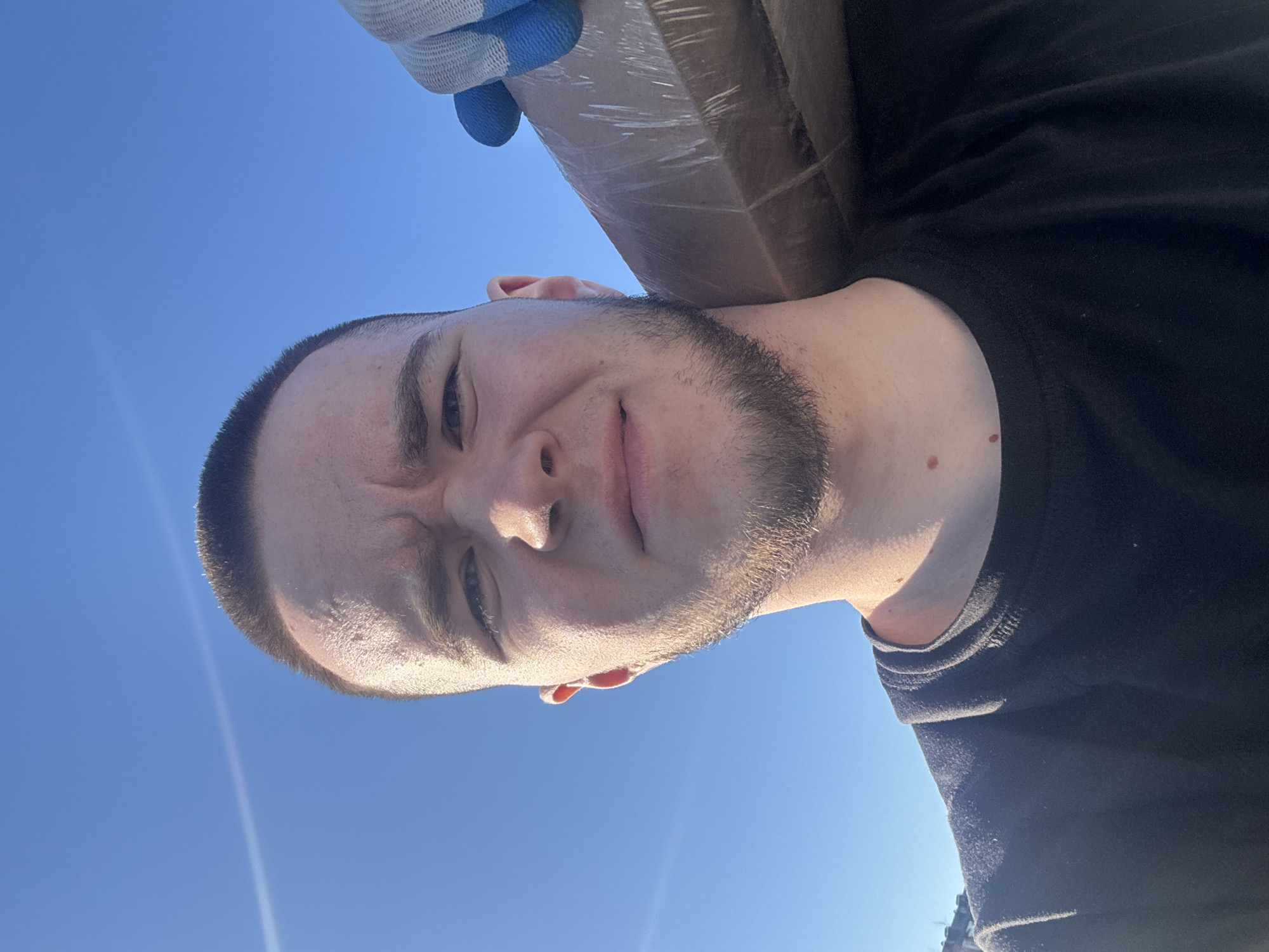 A medium shot of a man with a beard, dressed in a black T-shirt. He is holding a package wrapped in plastic on his shoulder with a gloved hand. The sky is clear and blue. The location is possibly Kazan, Russia, based on the presence of buildings on the horizon.