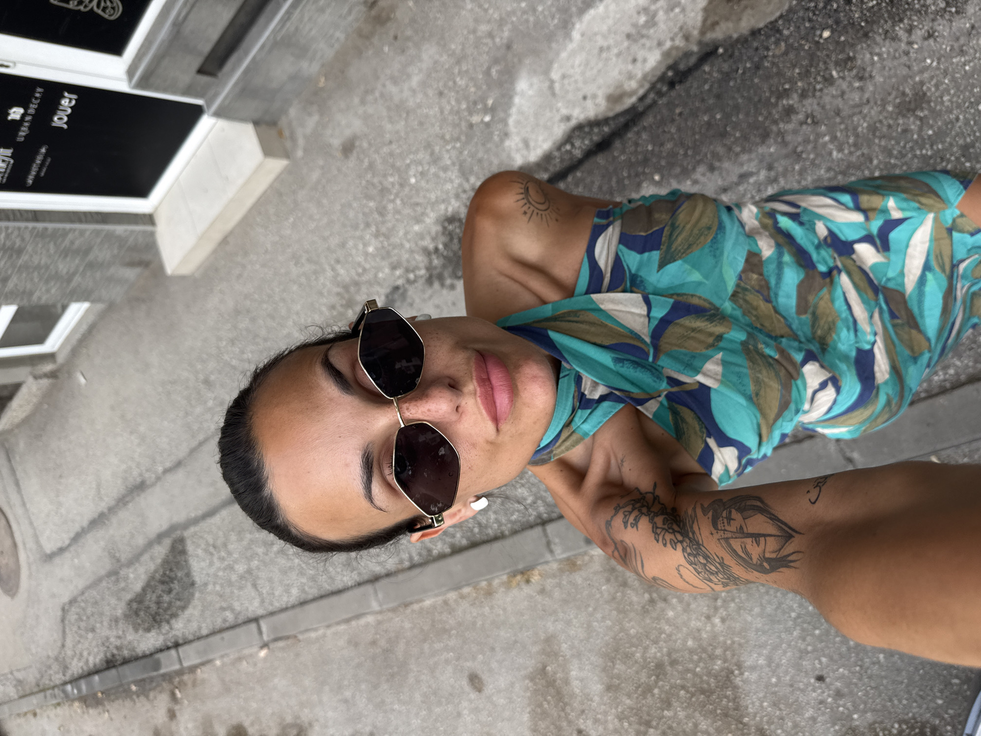 The image shows a person from a high-angle perspective, looking upwards towards the camera. The person is wearing dark-rimmed sunglasses and a blue and green patterned dress. They have tattoos on their arm and shoulder. The setting appears to be outdoors, on a paved surface that looks like a sidewalk or street. In the background, there is a building with a dark sign that has white text, and a window. The lighting suggests it is daytime. The person's expression is neutral or slightly amused. Visible text on the sign reads "URBAN DECAY" and "jouer". The image appears to be taken in Pleven, Bulgaria.