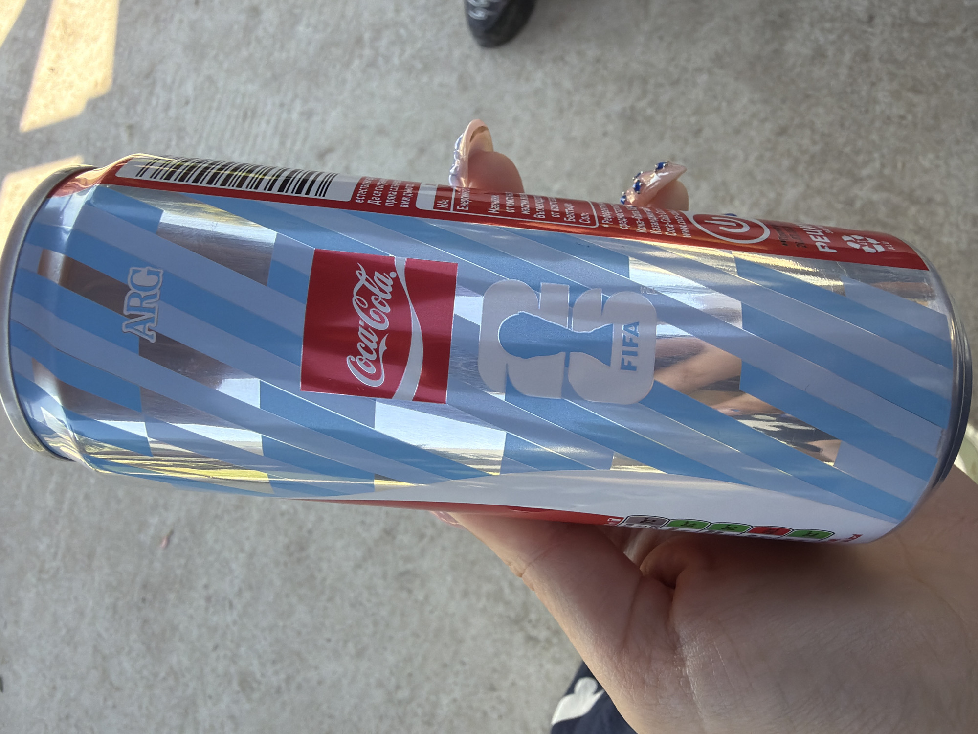 A hand with blue-jeweled fingernails holds a Coca-Cola can. The can is predominantly silver with light blue and white diagonal stripes. The Coca-Cola logo is in a red rectangle on the can, along with the word "FIFA." The can has text in a foreign language and a barcode. The background appears to be a light-colored concrete surface. The location is specified as Dinkata, Bulgaria.