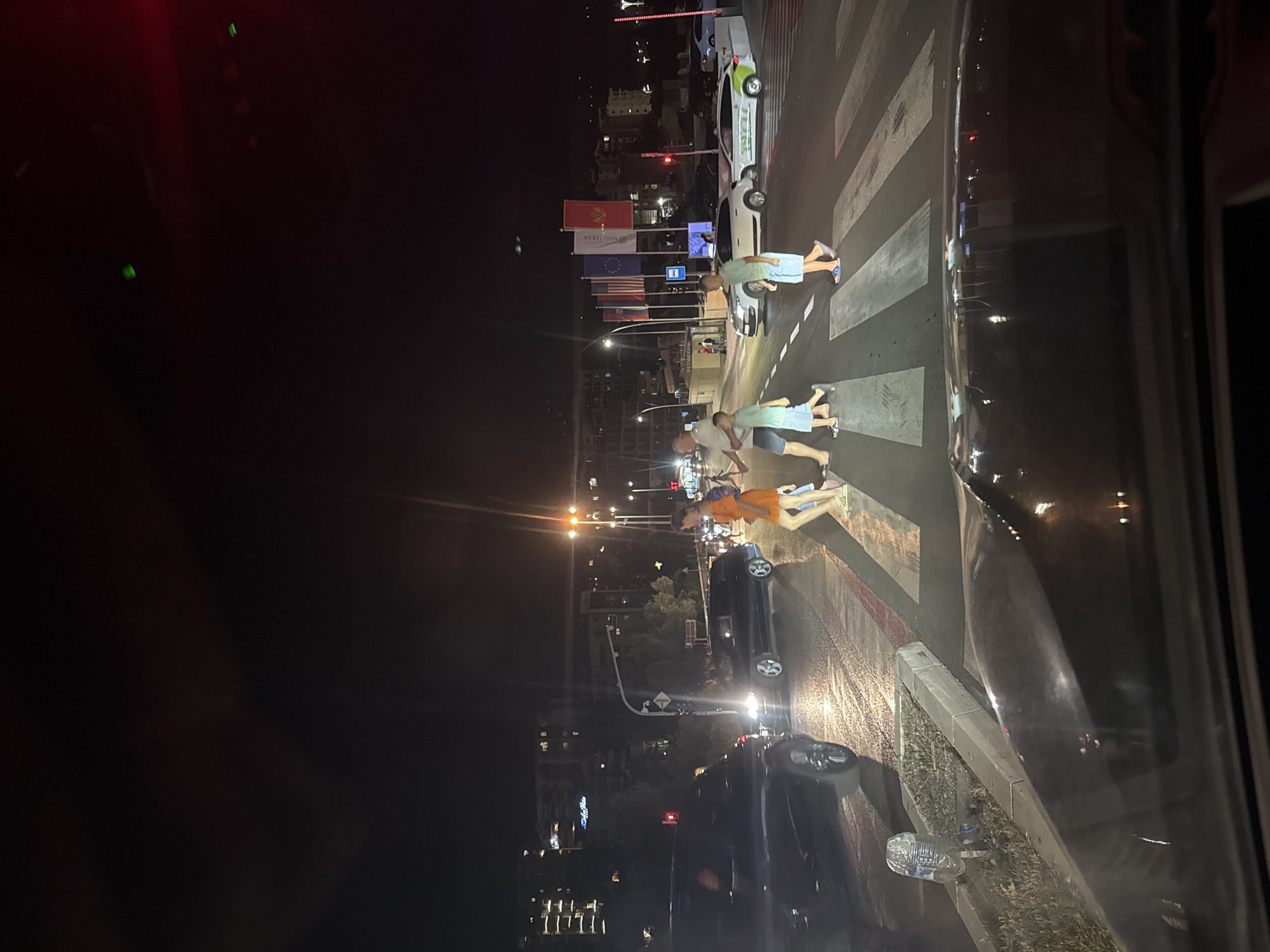 The image depicts a street scene at night, likely in the city of Boreti, Montenegro. A group of people, including adults and children, are crossing a marked pedestrian intersection. Several vehicles are visible, including dark-colored cars and a white vehicle with green and white markings. Buildings are illuminated in the background, suggesting an urban or resort area. The road surface appears wet, indicating recent rain or damp conditions. The scene is lit by streetlights and vehicle headlights, creating a dynamic play of light and shadow. Flags are displayed on poles in the background.