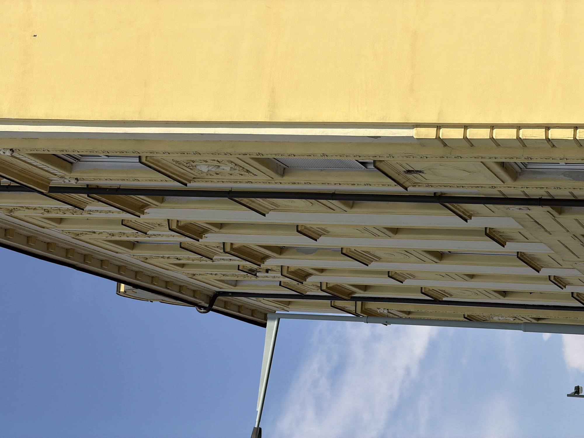 A side view of a pale yellow building with a blue sky in the background. The building has a classical architectural style, featuring multiple windows with decorative frames. A black downspout runs along the side of the building, and a light gray street lamp arm extends from the left side of the frame. The image was captured in Prague, Czechia.
