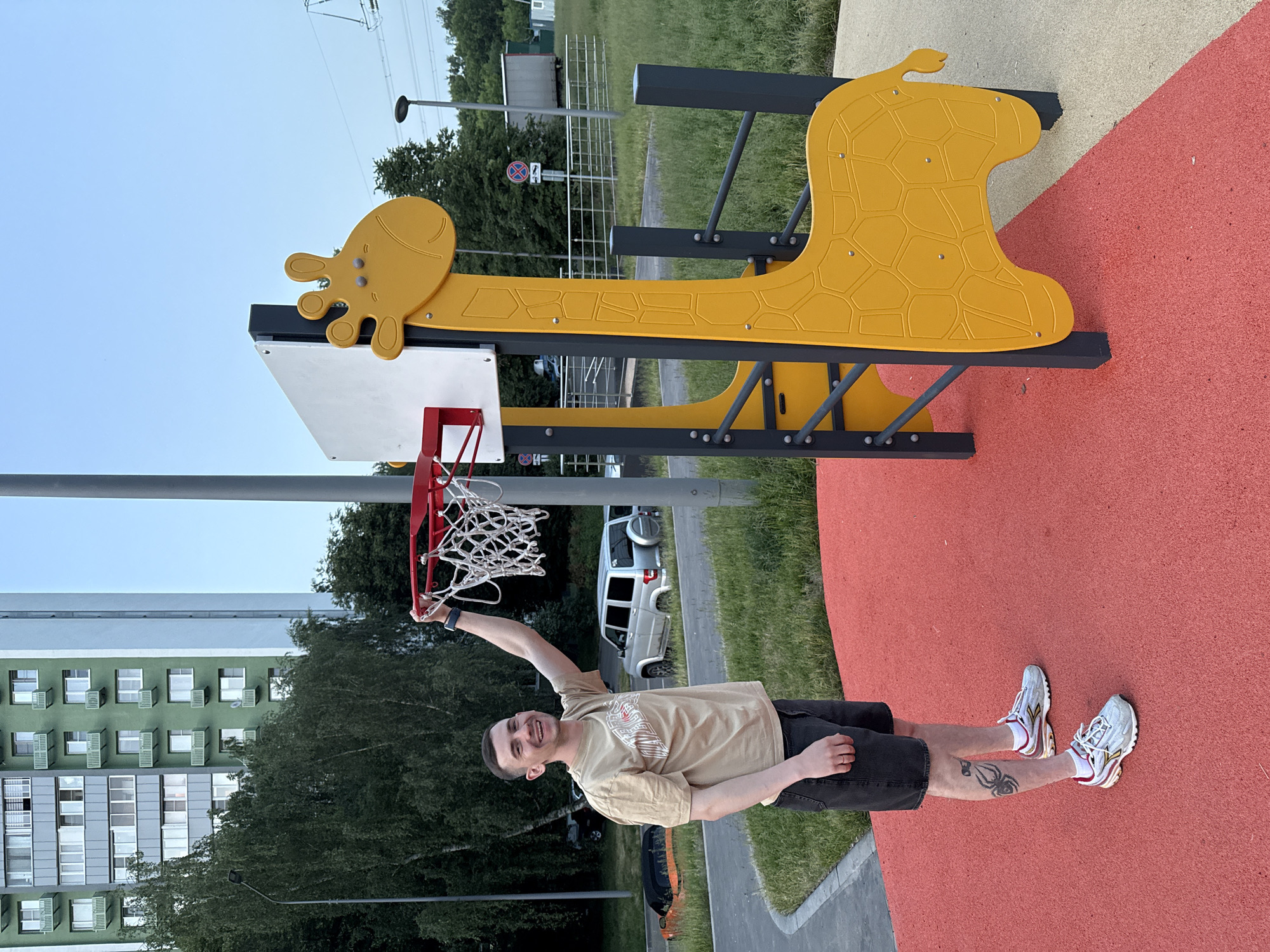 A young man is standing on a red rubberized playground surface, smiling and holding a basketball hoop with his right hand. He is wearing a tan t-shirt, dark shorts, white socks, and athletic shoes. He has a tattoo on his left thigh. Behind him, to the left, is a tall, green and white apartment building. To the right of the man is a children's playground structure designed like a yellow giraffe, with a white backboard attached. In the background, there are trees, grass, parked cars, and a street. The sky is clear and blue. The scene is outdoors during daylight hours. The location is on a playground in Moscow, Russia.