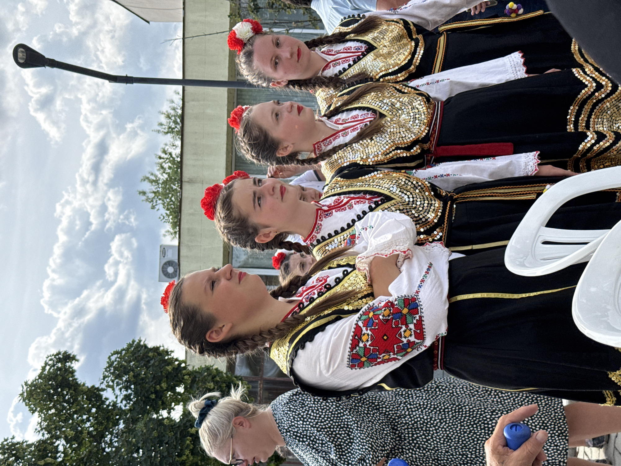In Pordim, Bulgaria, a group of four young women are pictured standing in a line, dressed in traditional Bulgarian folk costumes. The women are wearing white blouses with embroidered details on the sleeves, black skirts, and decorative vests adorned with gold sequins and trim. Each woman has red flowers in her braided hair. Behind them, a street lamp is visible, along with a building with an air conditioning unit and a tree. To the left of the women, an adult woman in a black and white patterned dress is partially visible, and a hand holding a blue object is in the lower left corner of the frame. The sky is cloudy.