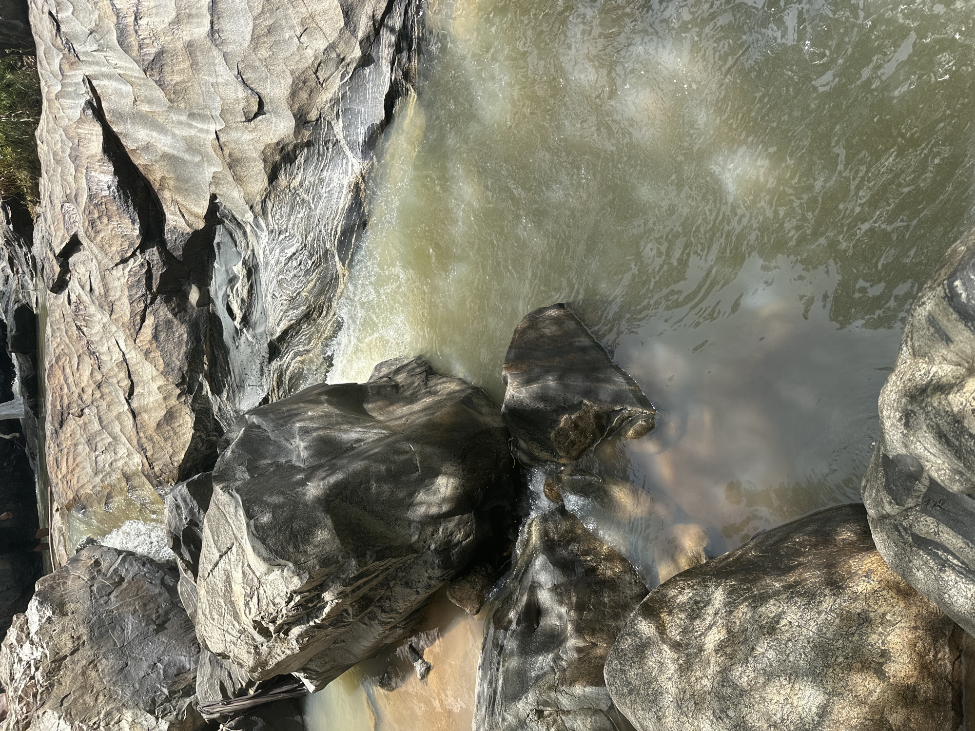The image shows a close-up view of a rocky riverbed with murky, light green water. Large, weathered rocks of various sizes and textures dominate the foreground and middle ground. Some rocks are smooth and rounded, while others have rough, layered surfaces with visible striations. Sunlight casts dappled shadows on the rocks and water. In the background, a small cascade of water flows over some of the rocks. A few indistinct figures can be seen standing in the water near the cascade, suggesting this is a location people visit. The environment appears to be natural and undeveloped. This scene is located in the city of Fobur, Nigeria.