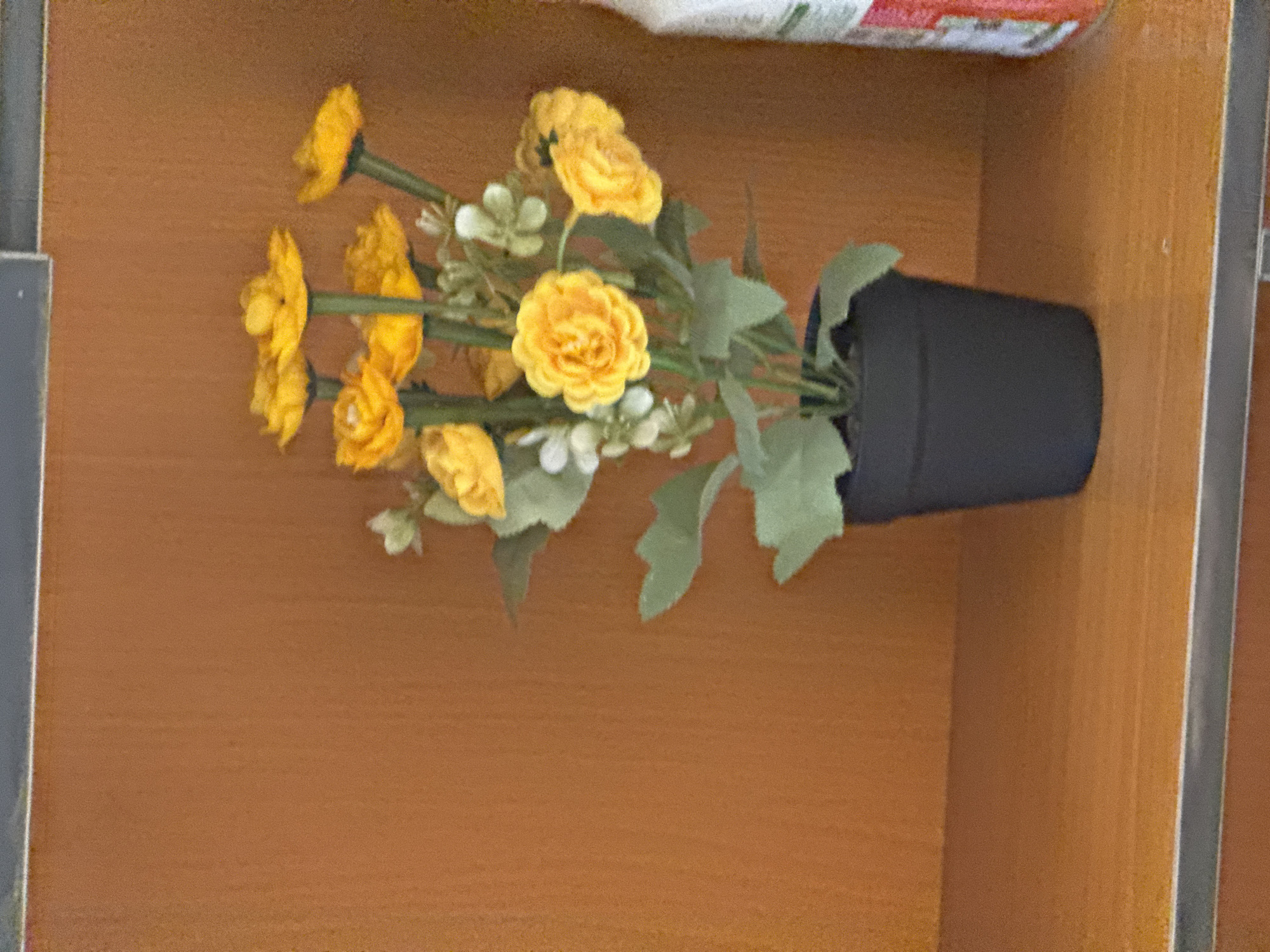 A flower arrangement with yellow flowers is in a black pot. The arrangement is sitting on a shelf in a wooden cabinet. The location is not identifiable as Abuja, Nigeria.