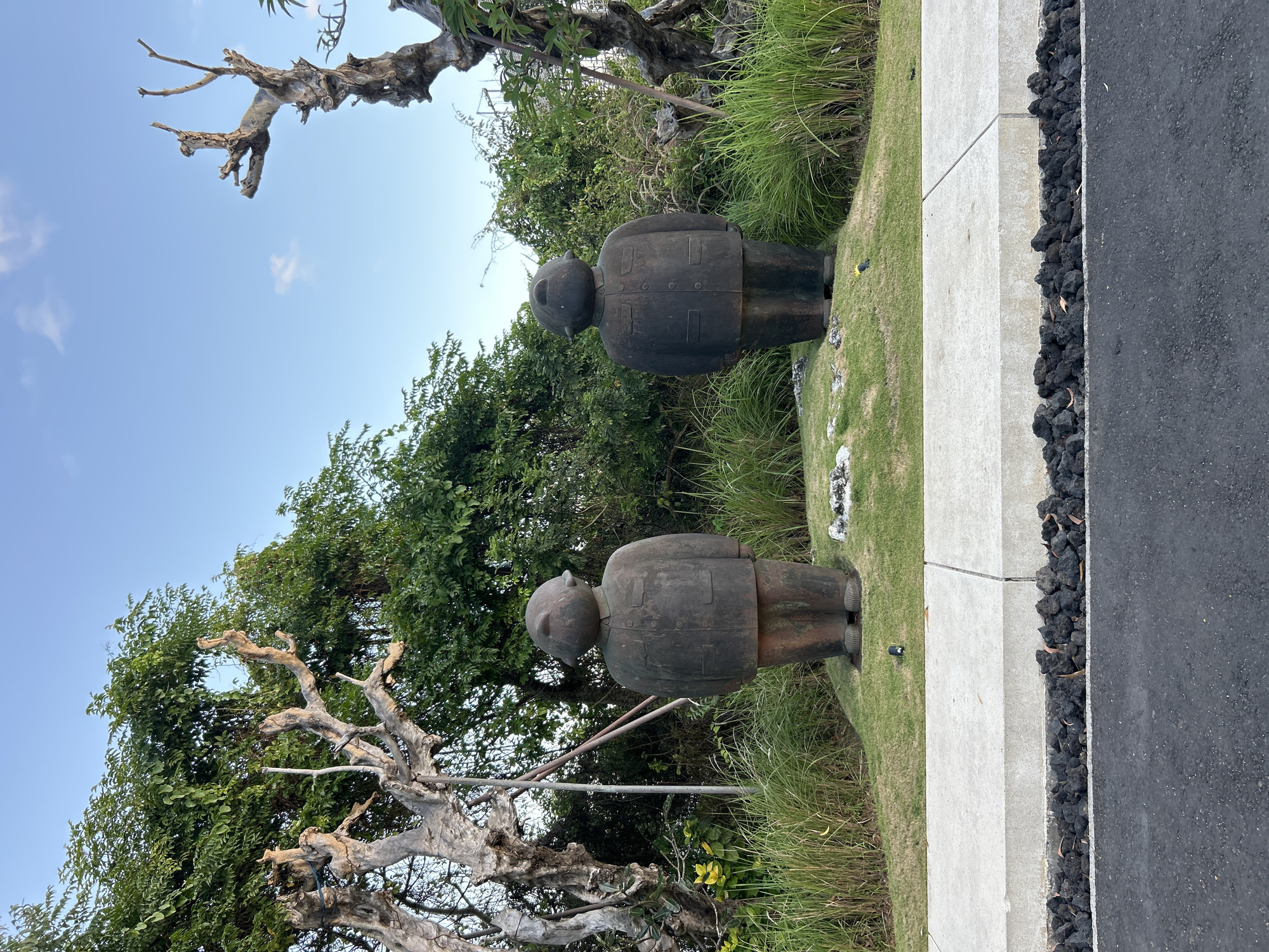 Two dark, weathered, roughly carved stone statues of people stand side-by-side on a grassy mound in Kuta Selatan, Indonesia. The statues are identical, appearing to be figures in jackets with small heads and rounded features. Behind them, dense green foliage and the bare, gnarled branches of dead trees create a textured backdrop against a clear blue sky. In the foreground, a grey concrete curb separates a paved road from the grassy area where the statues are situated, with dark volcanic rocks lining the base of the curb. The scene is illuminated by natural daylight, suggesting it was taken outdoors during the day.