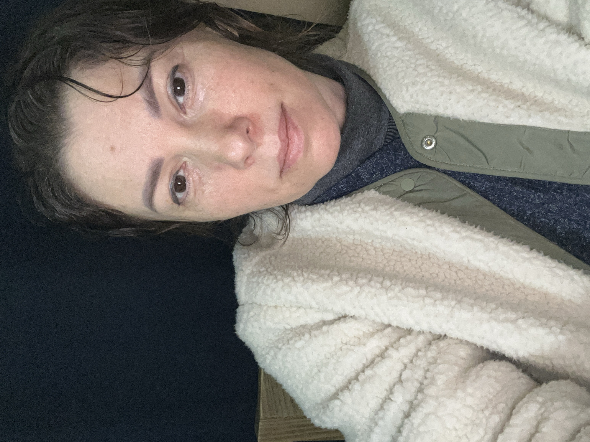 A woman with dark, damp hair pulled back from her face is looking directly at the camera. She has brown eyes and fair skin with a few freckles. She is wearing a white, fuzzy jacket over a gray turtleneck and a green outer garment. The background is a dark, plain color.