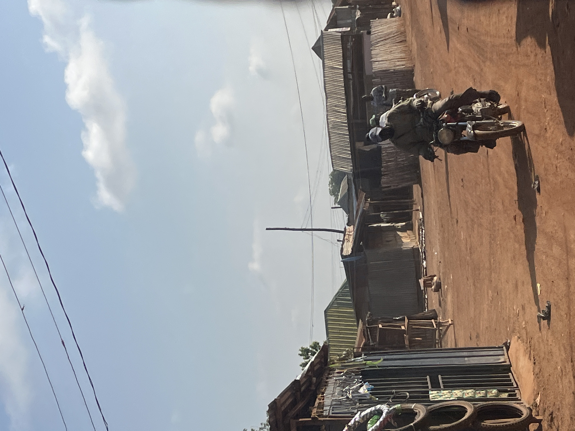 A motorcycle with two people is traveling on a dirt road, lined with shacks and basic buildings. The sky is visible with a few clouds. The environment appears to be a developing area. There is no explicit information to confirm if this is Abuja, Nigeria, or anywhere in Nigeria.