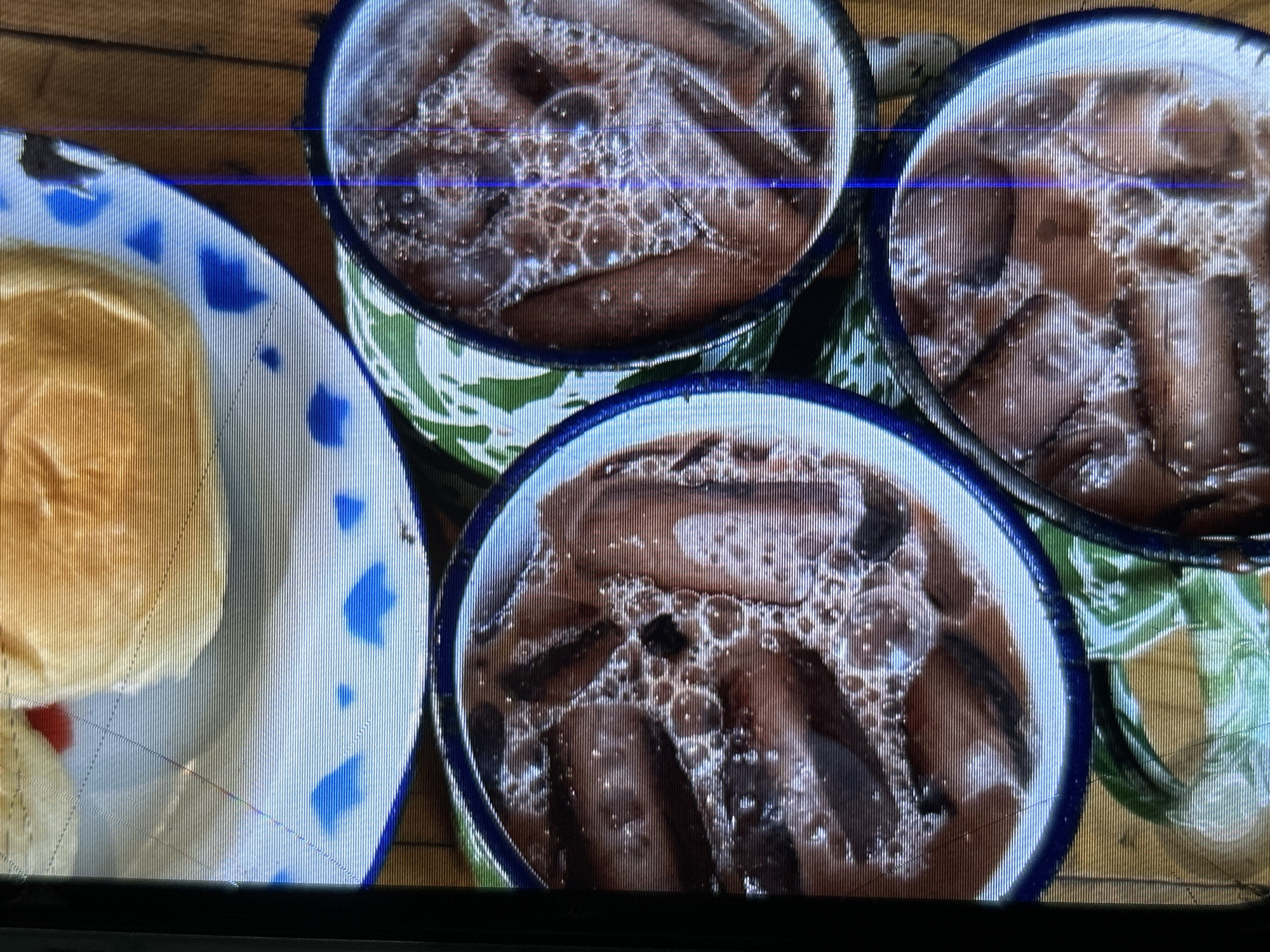 The image shows a close-up of three mugs filled with a dark brown beverage, likely iced chocolate milk, with ice cubes visible. The mugs have a blue rim and are placed on a surface with a green and white patterned placemat. In the background, on a white plate with blue patterns, is a bun with a red topping, possibly jam or fruit. The setting appears to be a cafe or restaurant, and the style of the mugs and plate suggests a rustic or traditional aesthetic. The image is taken from a high angle, looking down at the drinks. There is no discernible information within the image to indicate a specific location in Depok, Indonesia.