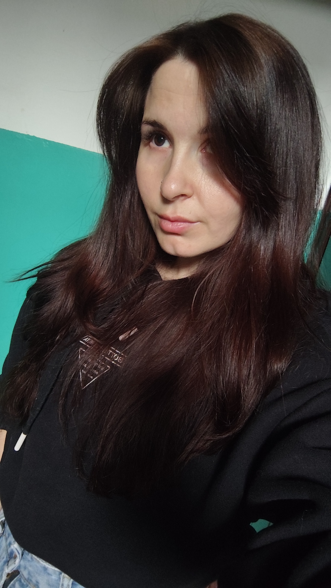 A young adult woman is featured in a head-and-shoulders portrait, likely a selfie, taken indoors in Фоки, Russia. She has long, voluminous dark brown or auburn hair that falls over her shoulders and chest. She looks slightly to the viewer's left with a neutral, calm expression, her lips gently parted. A small piercing is visible on her right nostril. She is wearing a dark, presumably black, hooded sweatshirt (hoodie). On the front of the hoodie, there is a light-colored graphic print consisting of a triangular logo. Partially legible text within the triangle includes "ATELIER" and a year, possibly "EST. 1980". Below her torso, light blue denim fabric is visible, suggesting she is wearing jeans or a similar garment. The background is an indoor wall, horizontally divided into two distinct colors: white on the upper section and a vibrant teal or turquoise on the lower section. The lighting is soft and even, typical of an indoor setting.