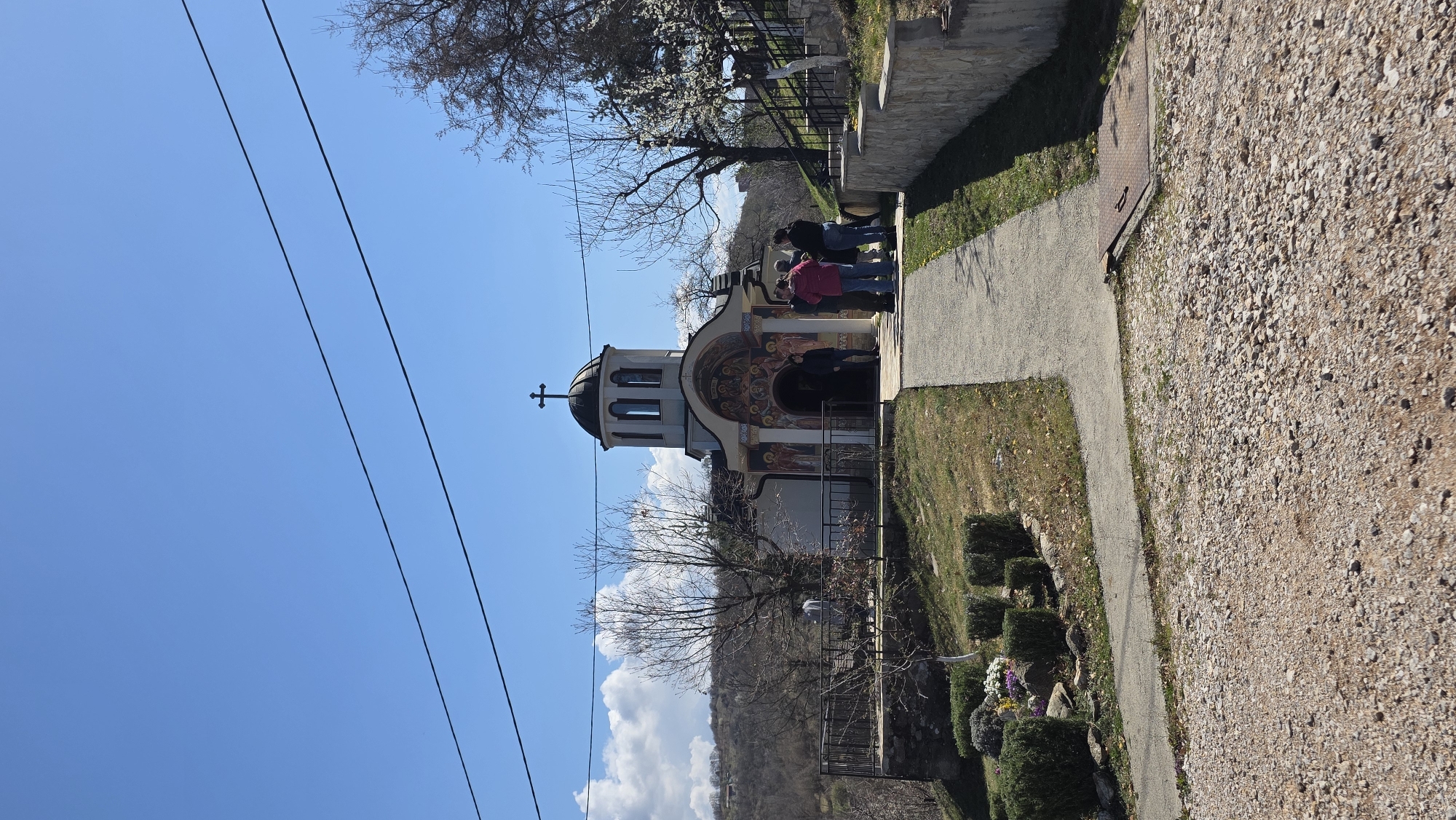 The image shows a small, ornate Eastern Orthodox church located in Elenovo, Bulgaria. A group of people is gathered at the entrance. The church has a dome topped with a cross, painted facades, and is set on a hillside. Power lines are visible against a clear blue sky with a few clouds. A paved path runs alongside the church, bordered by grass and small shrubs. A stone wall is further to the right of the path. The ground is covered with gravel.