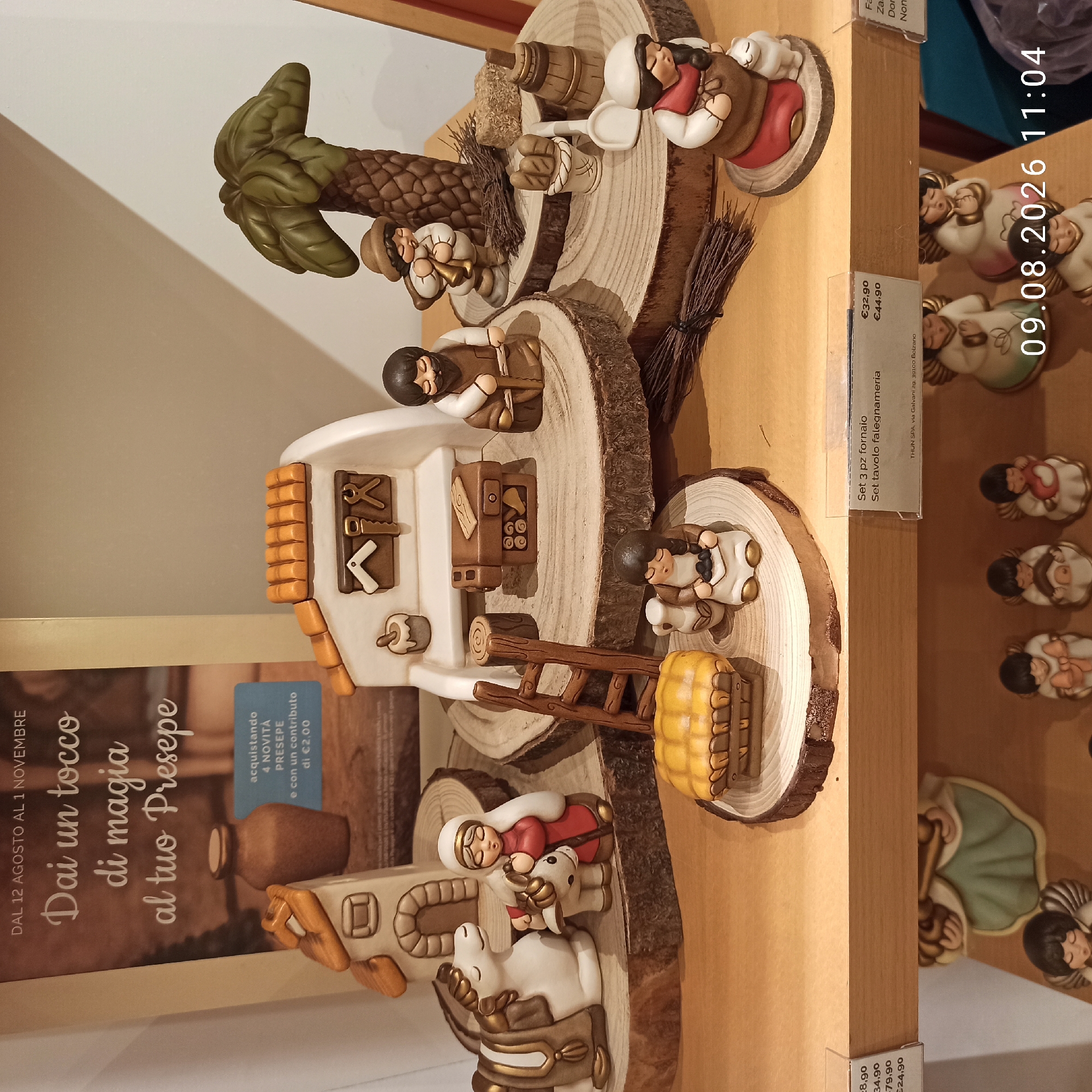 The image shows a display of ceramic Nativity figurines, likely in a shop or market. The figurines depict a Holy Family scene with Mary, Joseph, a donkey, and a stable. Other figures include a carpenter, a shepherd boy playing a horn, and a woman carrying a basket, all arranged on wooden slices. There is also a palm tree, a broom, hay bales, and bread. In the background, a poster reads "Dai un tocco di magia al tuo Presepe" (Add a touch of magic to your Nativity scene). Price tags are visible, with one indicating "Set 3 pz fornaio" (3-piece baker set) for €32.90 and "Set tavolo falegnameria" (workbench table set) for €44.90.
The timestamp "09.08.2026 11:04" is overlaid on the image.
The provided context of Molfetta, Italy is not visually verifiable from the image itself, as there are no landmarks or specific indicators within the scene to confirm this location. The scene is composed of manufactured objects for decorative purposes, arranged for display.