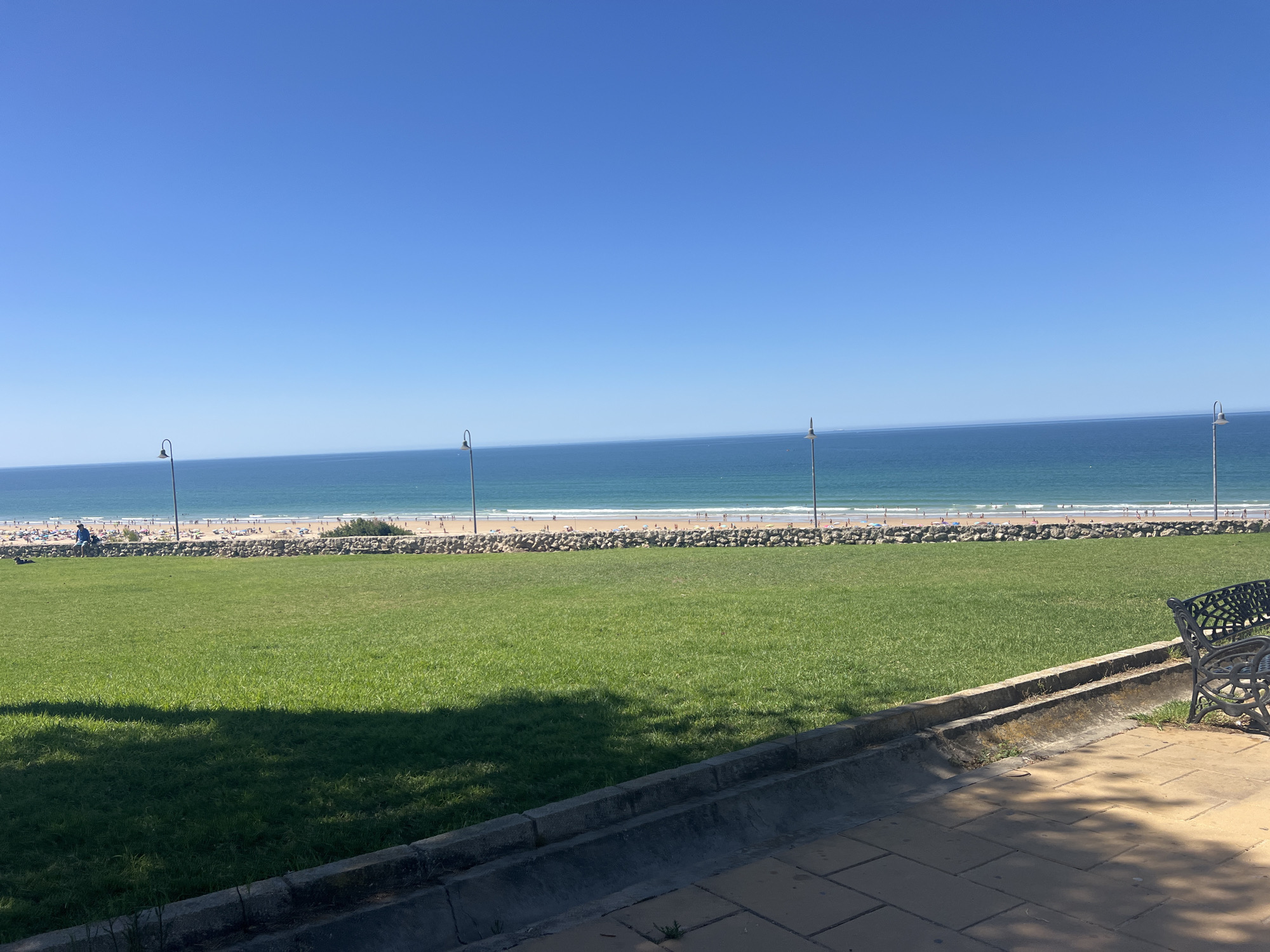 The image shows a serene view of a beach in Conil de la Frontera, Spain, on a clear, sunny day. The foreground features a well-maintained green lawn, with a portion shaded by trees. A stone-lined gutter runs along the edge of the lawn, and a decorative black metal bench with an ornate design is visible on the right side of the frame. Beyond the lawn, a stone wall separates the grassy area from the beach. The beach itself is populated with numerous sunbathers, and the turquoise waters of the ocean stretch out to the horizon, meeting a clear blue sky. Several lampposts are spaced along the beach area.