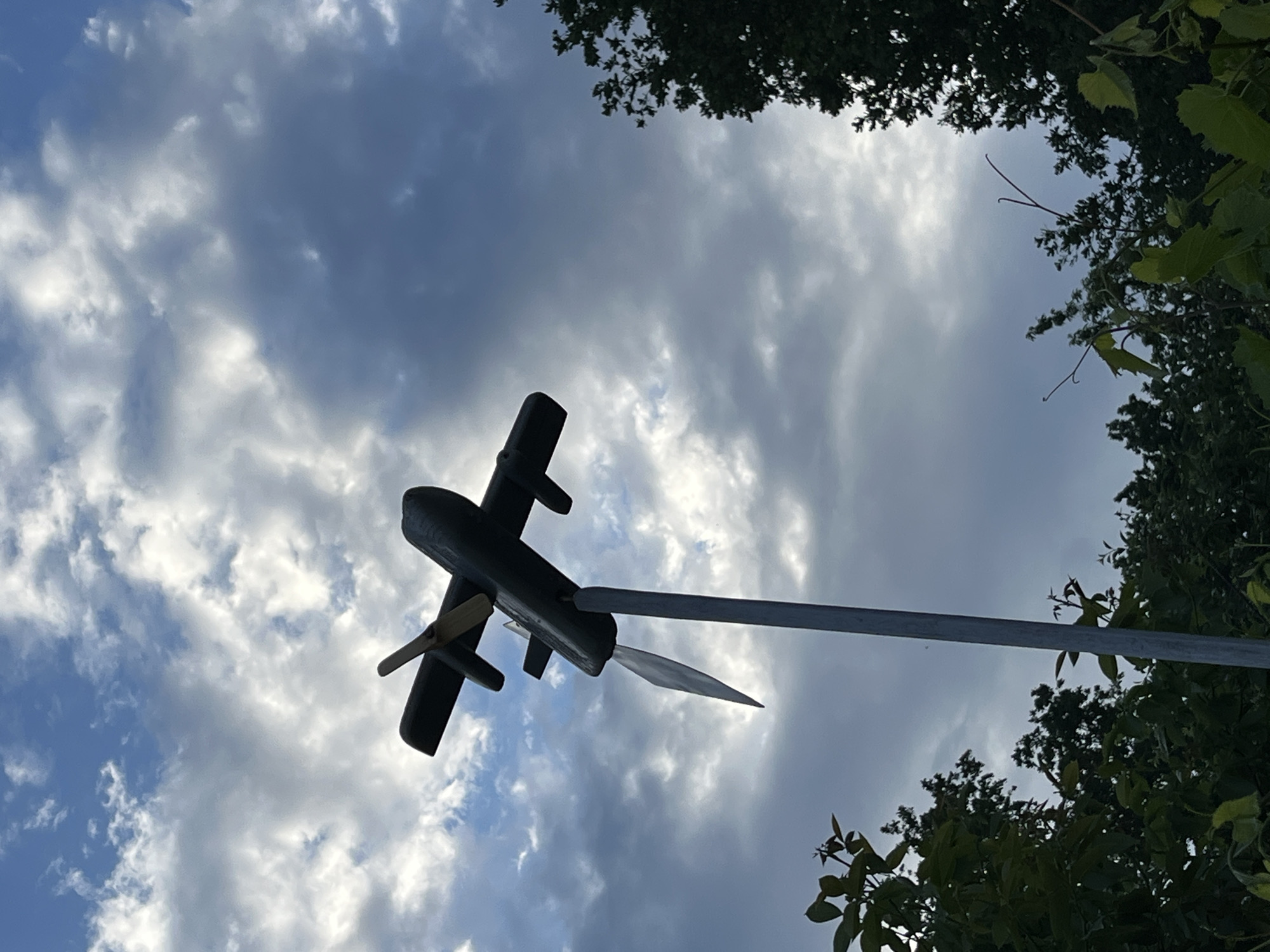 A wooden airplane weather vane is mounted on a tall metal pole. The airplane is dark-colored with a single wooden propeller at the front and two wings. A tail fin extends from the bottom of the airplane. The weather vane is positioned against a cloudy sky, with some trees visible in the foreground and along the bottom edge of the image.