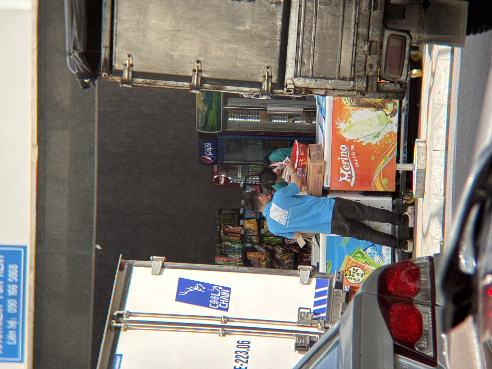 In an urban setting, possibly Nha Trang, Vietnam, a delivery truck is parked, its back doors open. A man in a blue t-shirt with a "Cool Chain" logo and dark pants is interacting with a refrigerator unit displaying ice cream advertisements. Next to him, a woman in a green shirt is also engaged with the products. Shelves stocked with various packaged goods are visible behind them. A car is parked in the foreground, partially obscuring the scene. The overall atmosphere suggests a commercial exchange or delivery occurring on a street.
