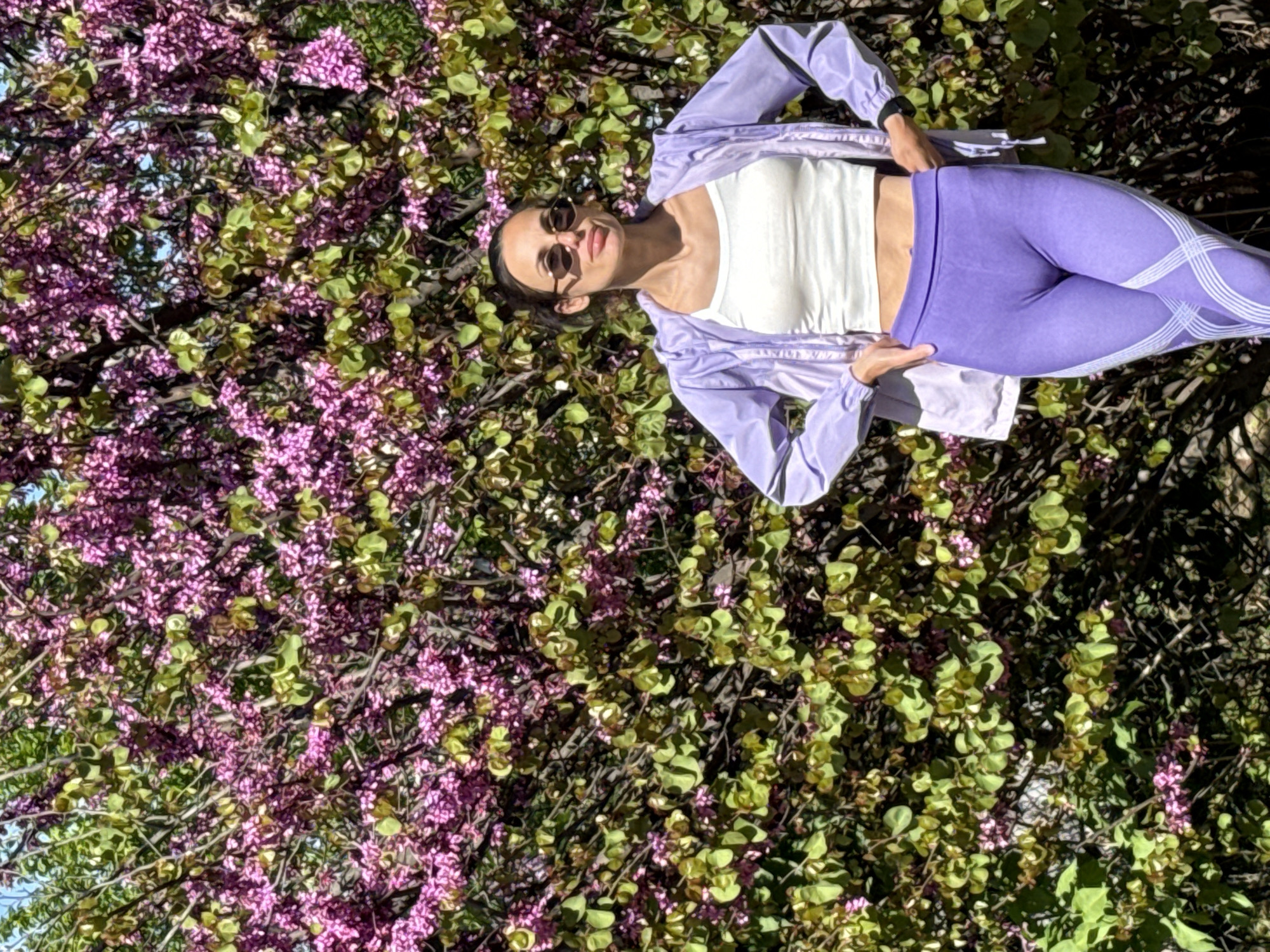 The original image is rotated 90 degrees clockwise, depicting a young woman in a full-body shot, appearing to lie amidst or recline against a dense backdrop of flowering trees. She wears dark sunglasses and has her hair pulled back. Her outfit consists of a white cropped tank top, a light lavender or purple zip-up jacket worn open, and matching purple leggings featuring distinctive white criss-cross patterns along the sides. A dark watch or fitness tracker is visible on her left wrist. She looks towards the camera with a relaxed expression and a faint smile.
The scene is dominated by abundant natural foliage, primarily a flowering tree, possibly a Judas tree, characterized by numerous small, dense pink and purple blossoms covering its branches. Bright green leaves are interspersed among the flowers. Small glimpses of blue sky can be seen through the foliage. The image is brightly illuminated by natural sunlight, indicating a clear day, likely in spring, given the blooming flowers. The location context is Pleven, Bulgaria. There is no visible text in the image.