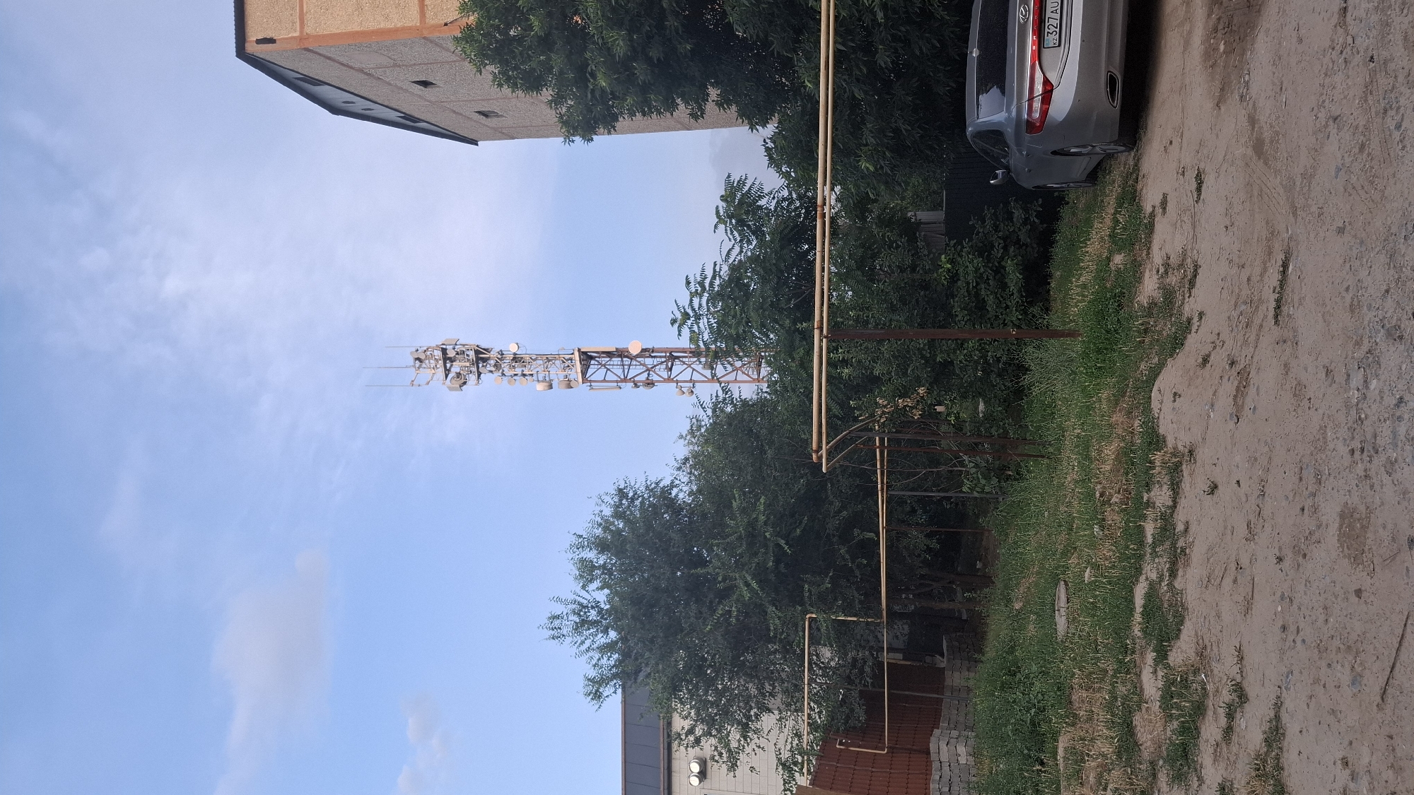 The image shows an outdoor scene on a sunny day, likely in Shymkent, Kazakhstan. The primary focus is a tall, metal telecommunications tower covered in various antennas and dishes, positioned against a clear blue sky with scattered clouds. The tower is set amidst lush green trees and foliage.
To the left of the tower, a portion of a multi-story building with a light-colored facade and dark-framed windows is visible. On the right side of the image, a light-colored car is parked on a dirt road. The car has a license plate with text that appears to be "KZ 327 AU". Some fencing and pipes are also present in the foreground and mid-ground. The overall setting suggests an urban or suburban environment where infrastructure and nature coexist. The lighting indicates it is daytime. There are no people visible in the image.