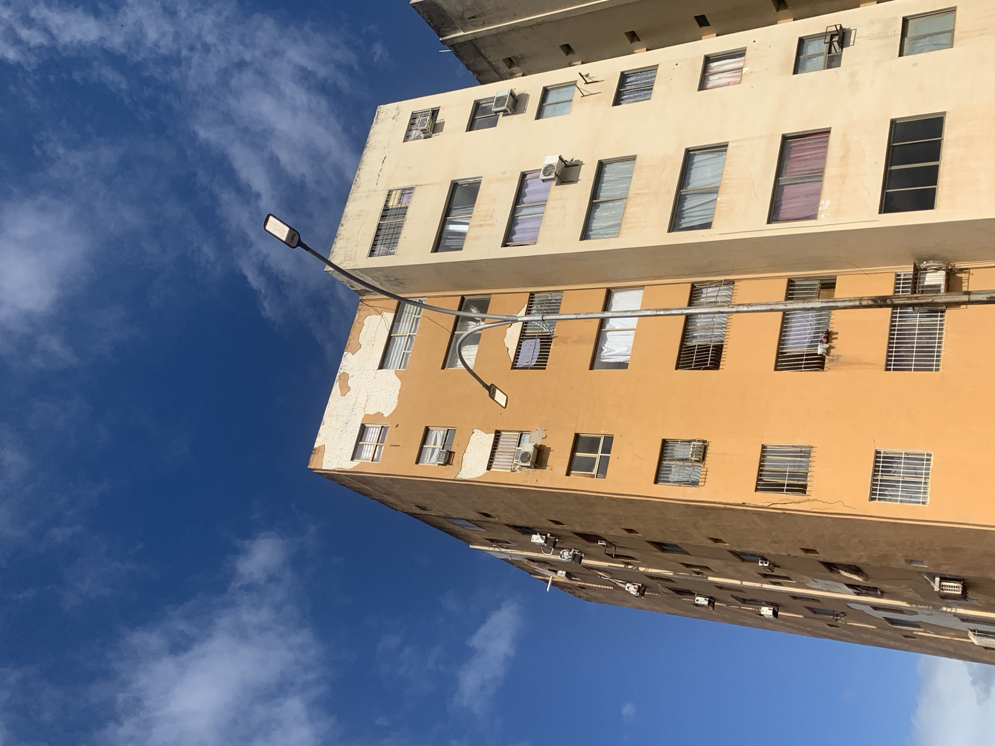 A low-angle shot captures two adjacent, tall apartment buildings against a vibrant blue sky with scattered white clouds. The building on the left is painted a bright orange and exhibits peeling paint on its upper section. It has numerous windows, some with white blinds, others with bars, and several air conditioning units mounted on the exterior. The building on the right is painted a pale yellow and also features many windows, some with curtains. A modern street lamp with a rectangular LED fixture stands prominently in the foreground, extending upwards between the two buildings. The scene appears to be in a city environment.