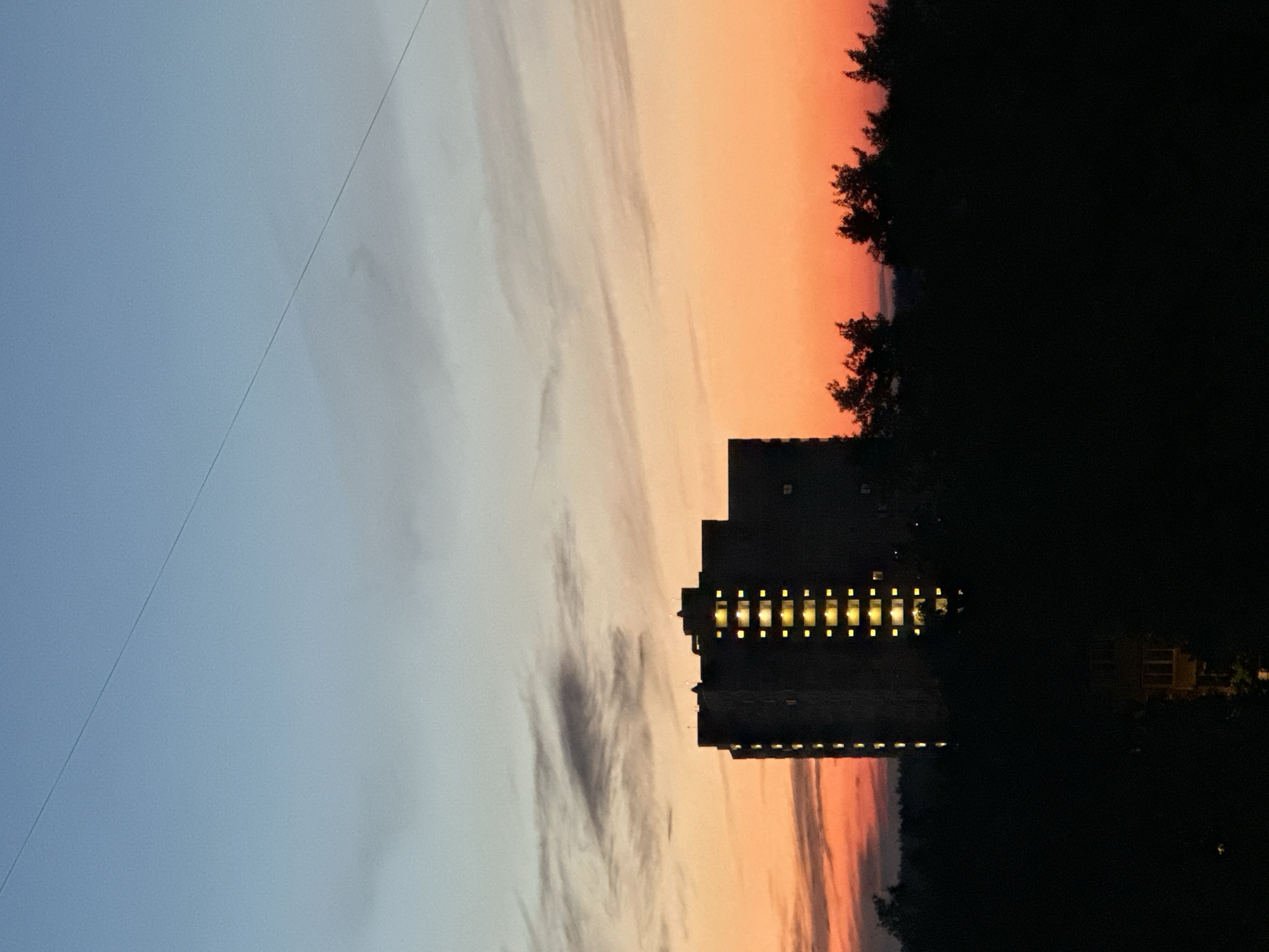 The image shows a dark silhouette of a multi-story apartment building against a twilight sky. The sky transitions from a pale blue at the top to a vibrant orange and pink near the horizon, with wispy clouds scattered throughout. Lights are visible in the windows of the building, suggesting that people are inside. The foreground is dominated by the dark shapes of trees, obscuring the ground level of the scene. A thin wire runs diagonally across the upper portion of the sky. The overall impression is one of a quiet evening in an urban or suburban setting.